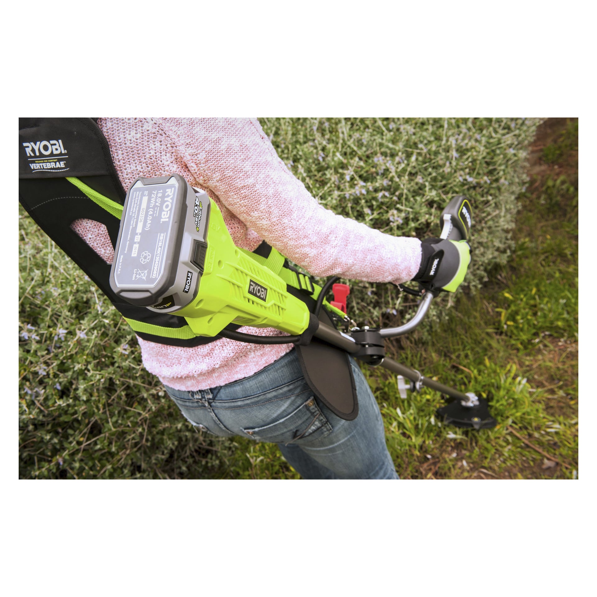 En person klädd i jeans och rosa tröja trimmar gräs och buskar i en trädgård med hjälp av Ryobi One+ OBC1820B batteridriven borstklippare med fastsatt sele.