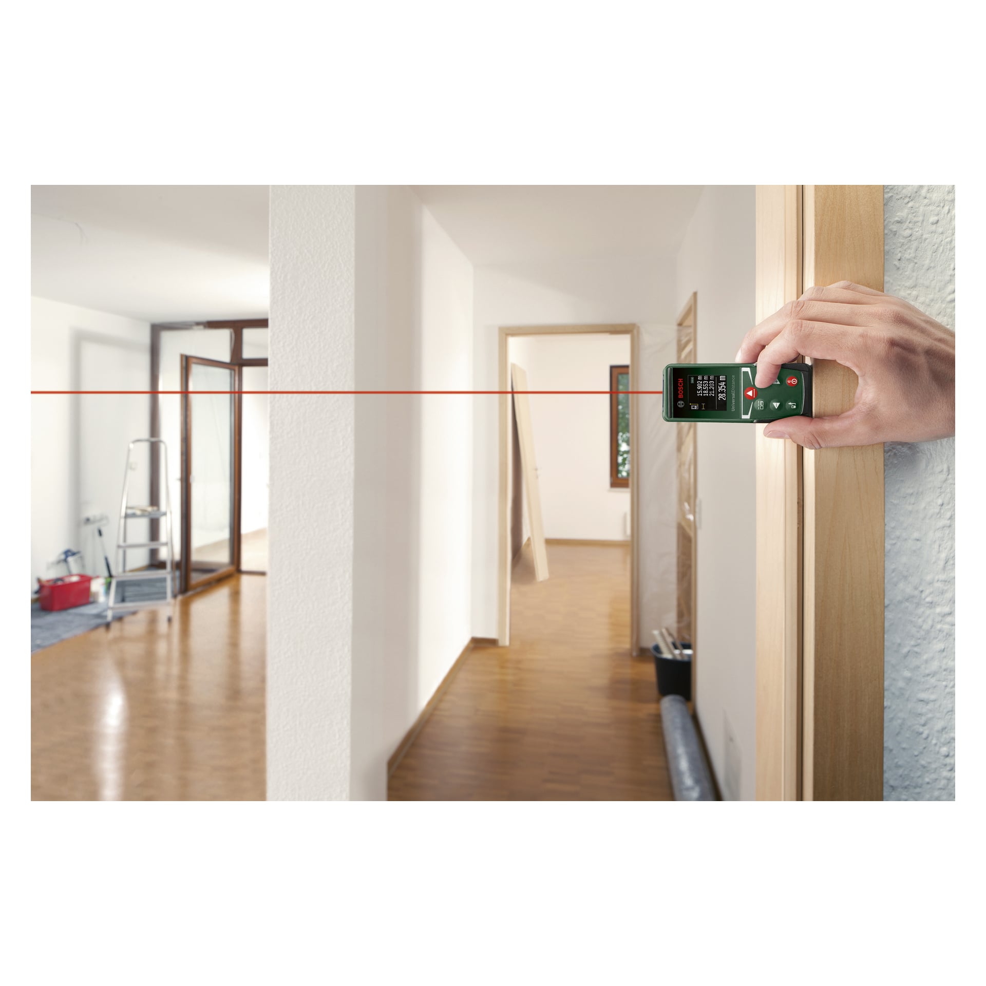Ręka trzyma miernik odległości Bosch UniversalDistance 30 przy drzwiach, a czerwony promień laserowy rozciąga się po drewnianej podłodze korytarza. W następnym pomieszczeniu widoczna jest drabina i kilka narzędzi.