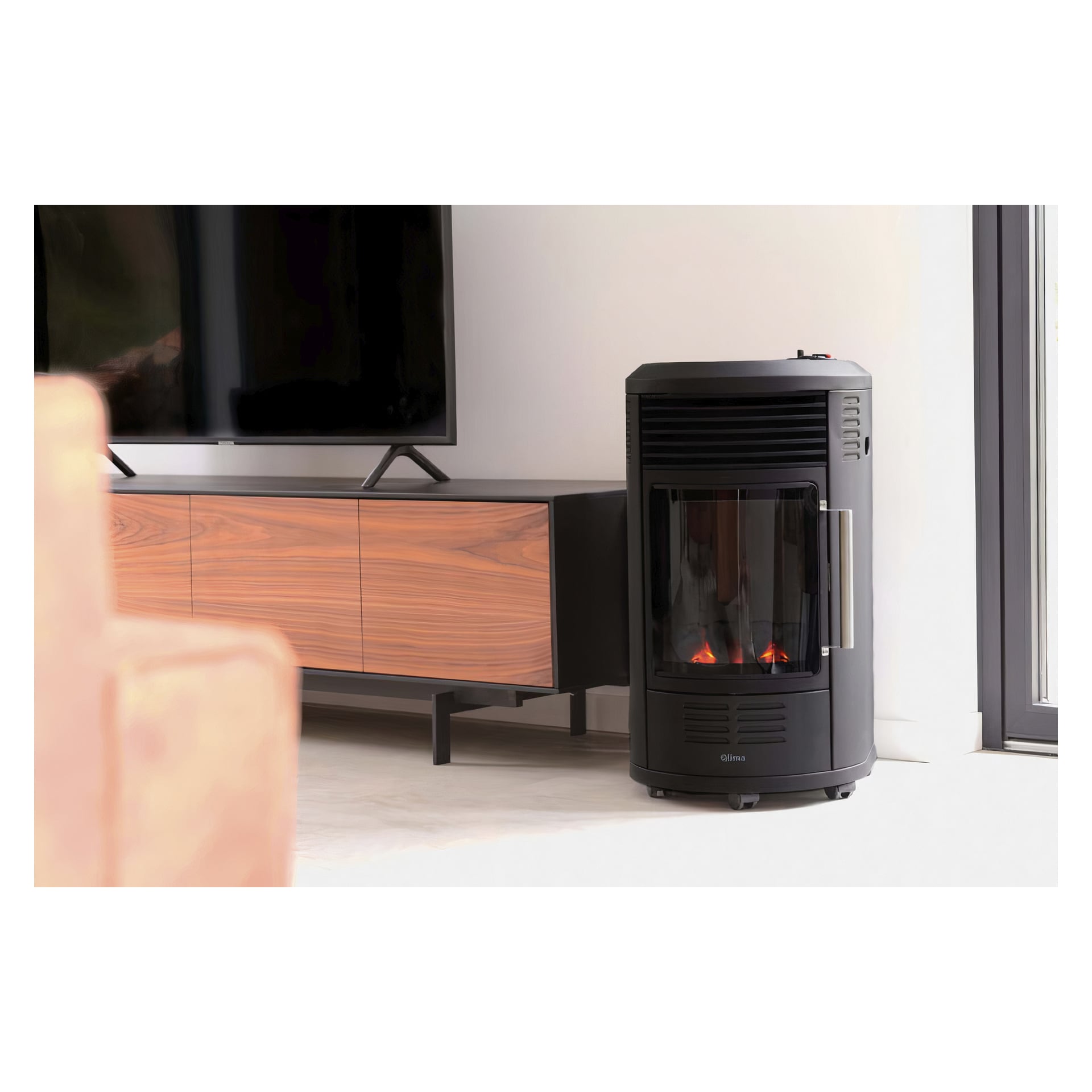 Qlima LPG-ovnen GH8034, en slank svart ovn med synlige flammer, står nær et treskap og en TV i en moderne stue med lyse vegger og gulv.