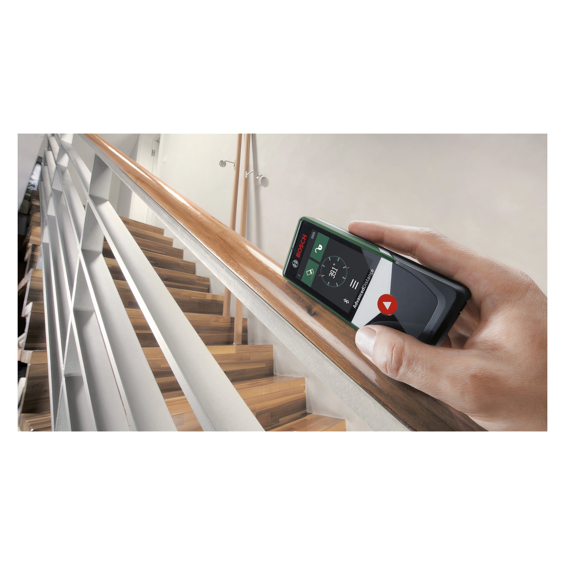 Käsi pitää Bosch AdvancedDistance 50 C -etäisyysmittaria puista portaikkoa vasten, jonka näytöllä näkyy mitat ja numerot lainausmerkeissä. Taustalla on moderni portaikko, jossa on puiset portaat ja valkoinen seinä.