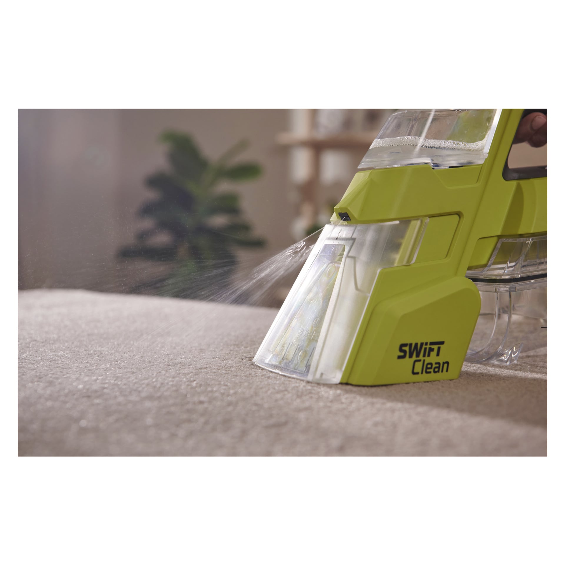 Zbliżenie na bateriowy odplamiacz Ryobi One+ Swift Clean RDC18-0, który rozprowadza płyn na beżowy dywan wewnątrz, z delikatnie rozmytym rośliną i półką w tle.