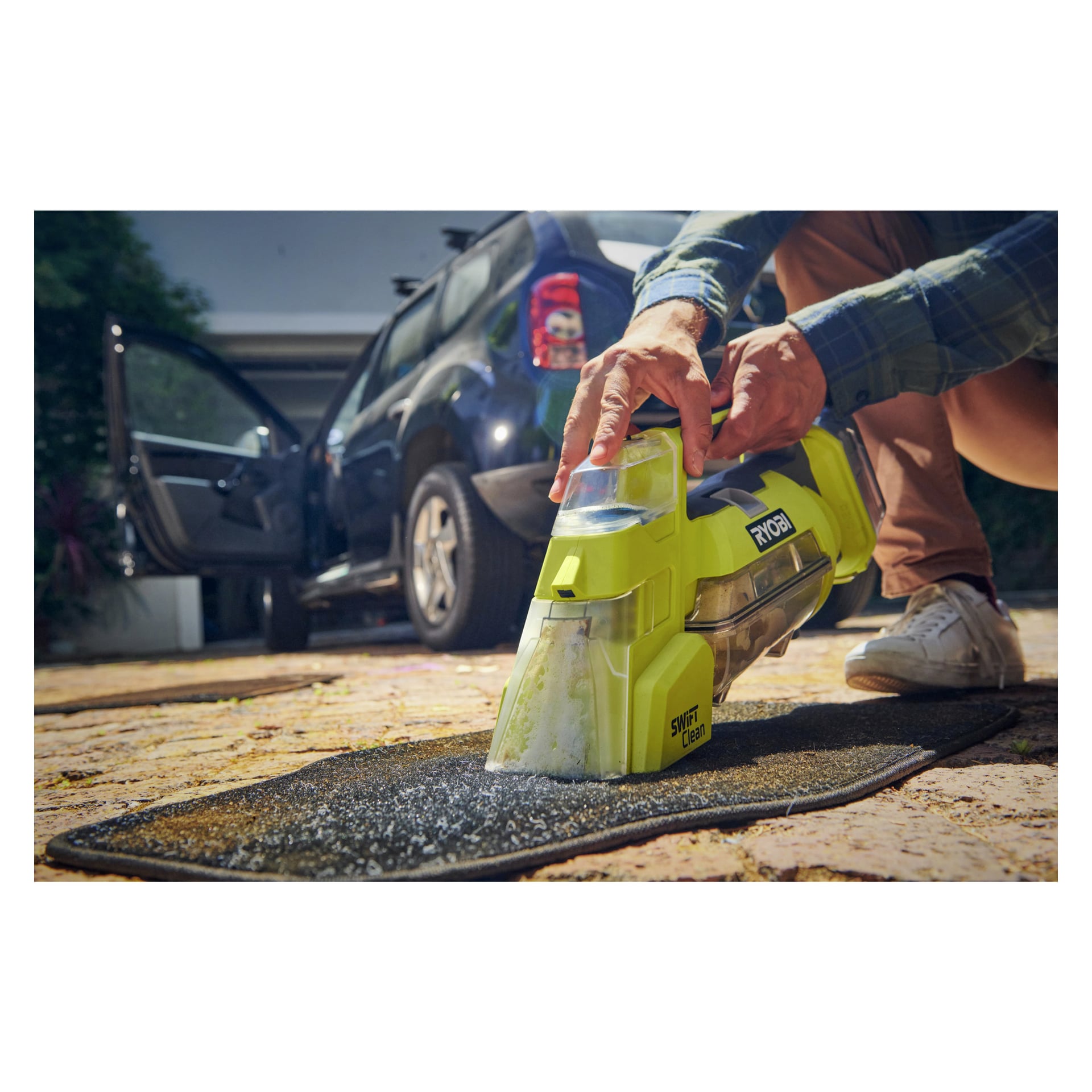 Ktoś obsługuje odplamiacz do baterii Ryobi One+ Swift Clean RDC18-0 na czarnym dywaniku samochodowym na zewnątrz, podczas gdy w tle widać niebieski samochód z otwartymi drzwiami.