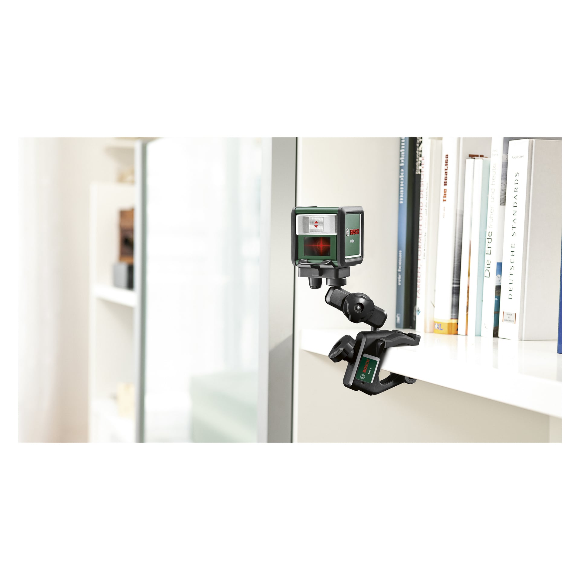 En Bosch Quigo III korslaser är fäst vid kanten av en hylla och gjuter ett rött laserkors på en upprätt glasyta. I bakgrunden finns böcker och extra hyllor.