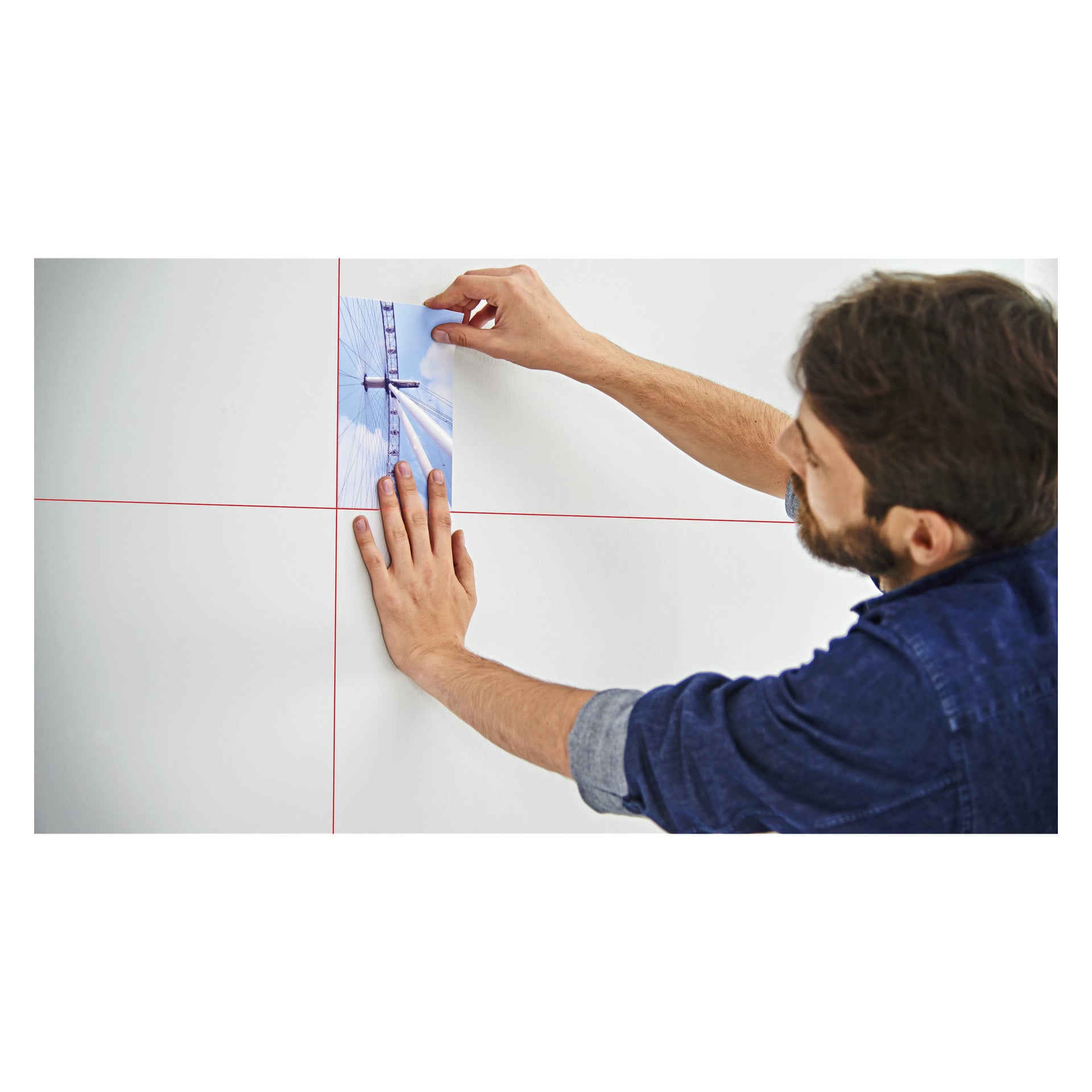 En man manövrerar Bosch Cross Laser Quigo III och projicerar röda korsande laserlinjer medan han placerar ett litet foto av en hängbro på en vit vägg.
