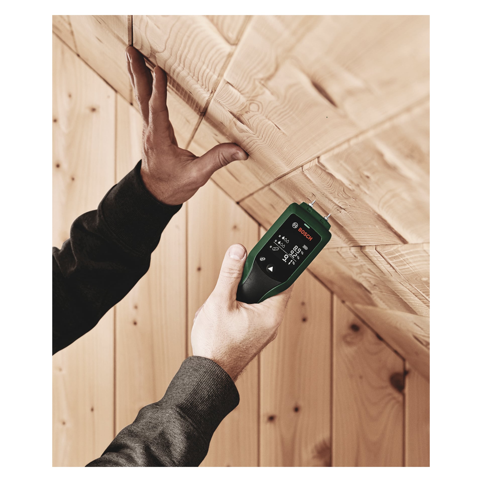 En person kontrollerar en träpanels i taket med en grön Bosch UniversalHumid handhållen digital fuktmätare, som visar mått på skärmen.