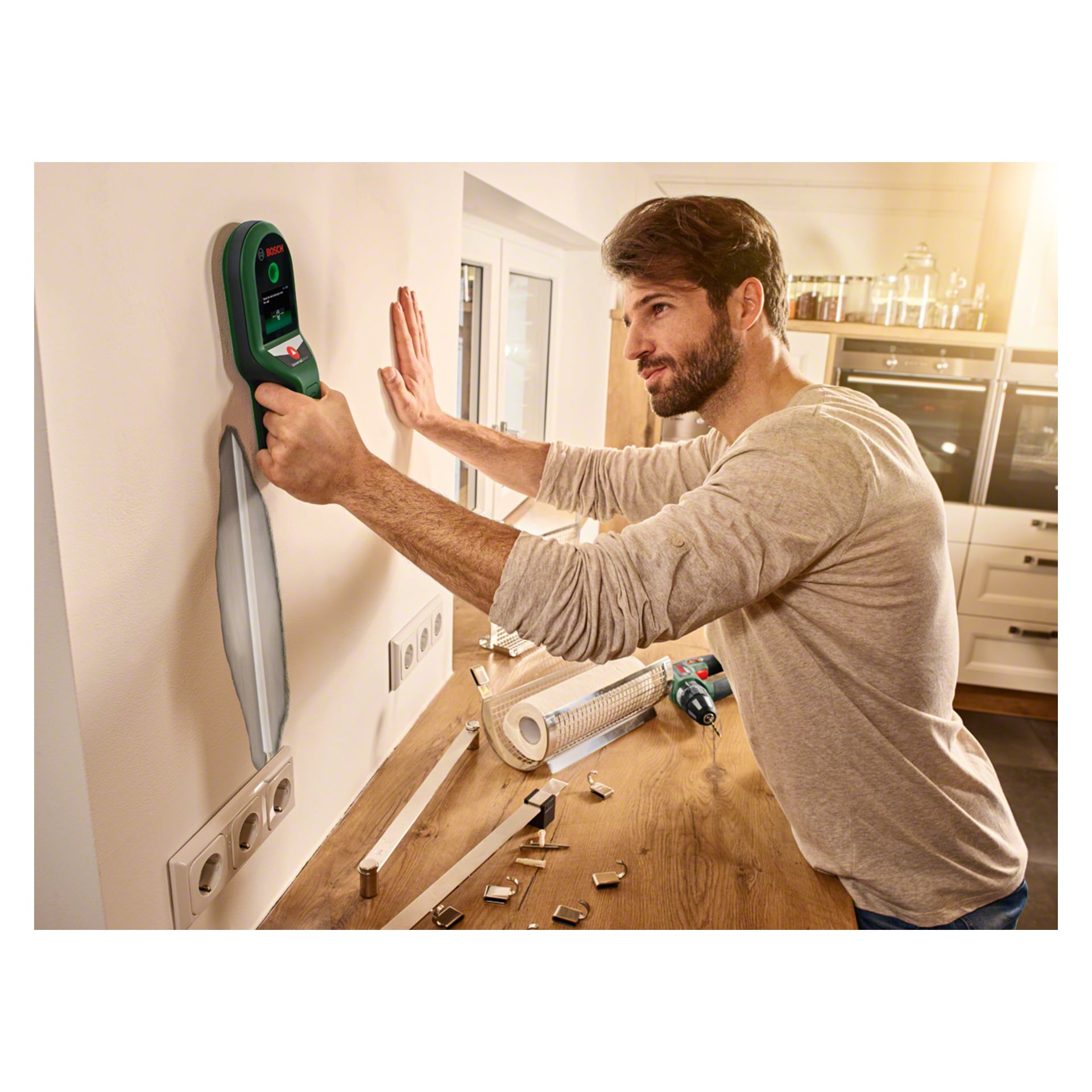 En man använder Bosch Multi-detektor UniversalDetect mot en vit vägg i ett elegant kök. Olika verktyg och beslag är utspridda på träbänken, med en sladdlös borr placerad i närheten. Han koncentrerar sig intensivt på enhetens display medan han arbetar.