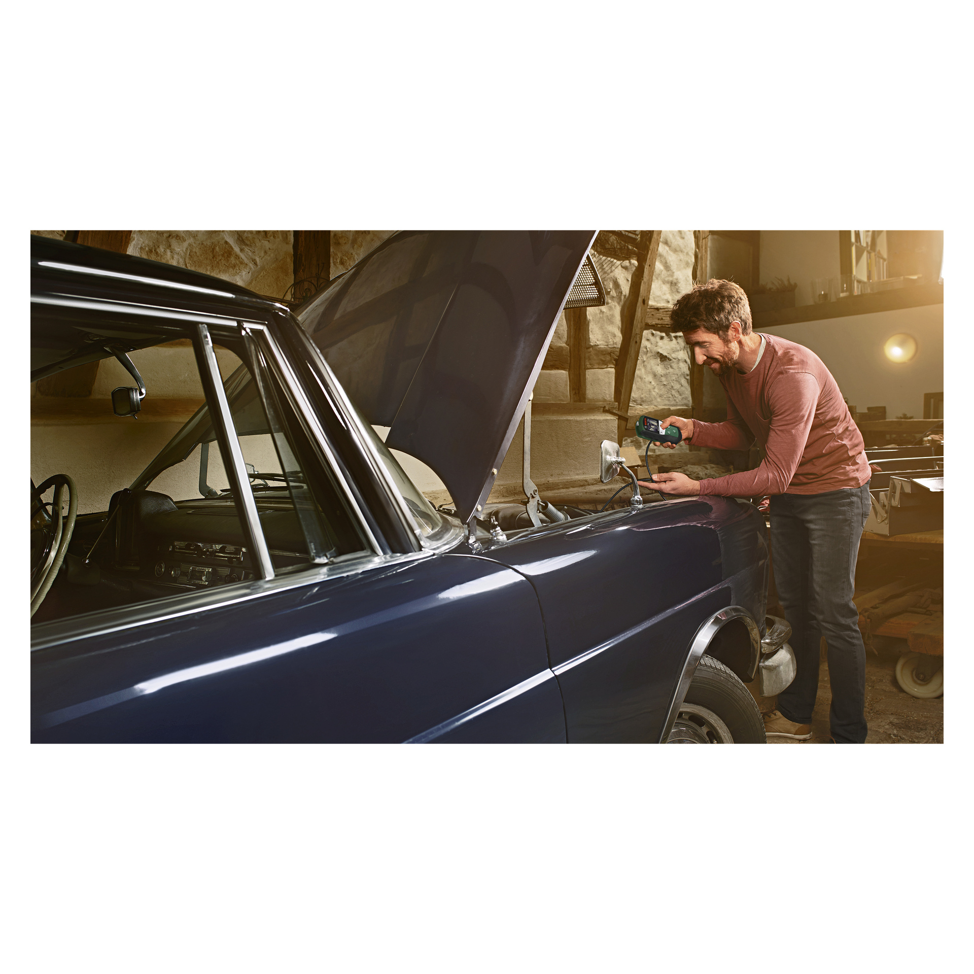 I ett garage står en man bredvid en klassisk blå bil med huven uppdragen, och använder Bosch UniversalInspect-inspektionskameran för att kontrollera eller reparera fordonet medan solljuset strömmar in genom ett fönster bakom honom.