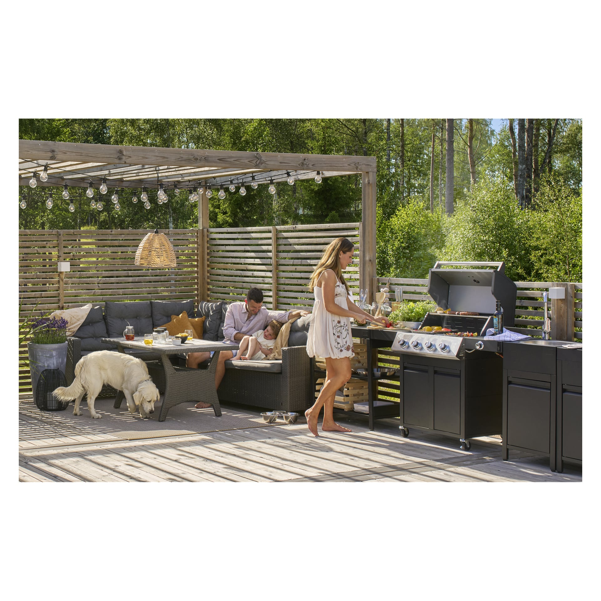 På en uteplats omgiven av trästaket och frodig grönska lagar en kvinna mat på en Burns & Barkles Gas Grill Steely King 6342B. I närheten ligger en man och ett barn på en utomhussoffa medan en vit hund utforskar altanen under lysande ljusslingor ovanför.