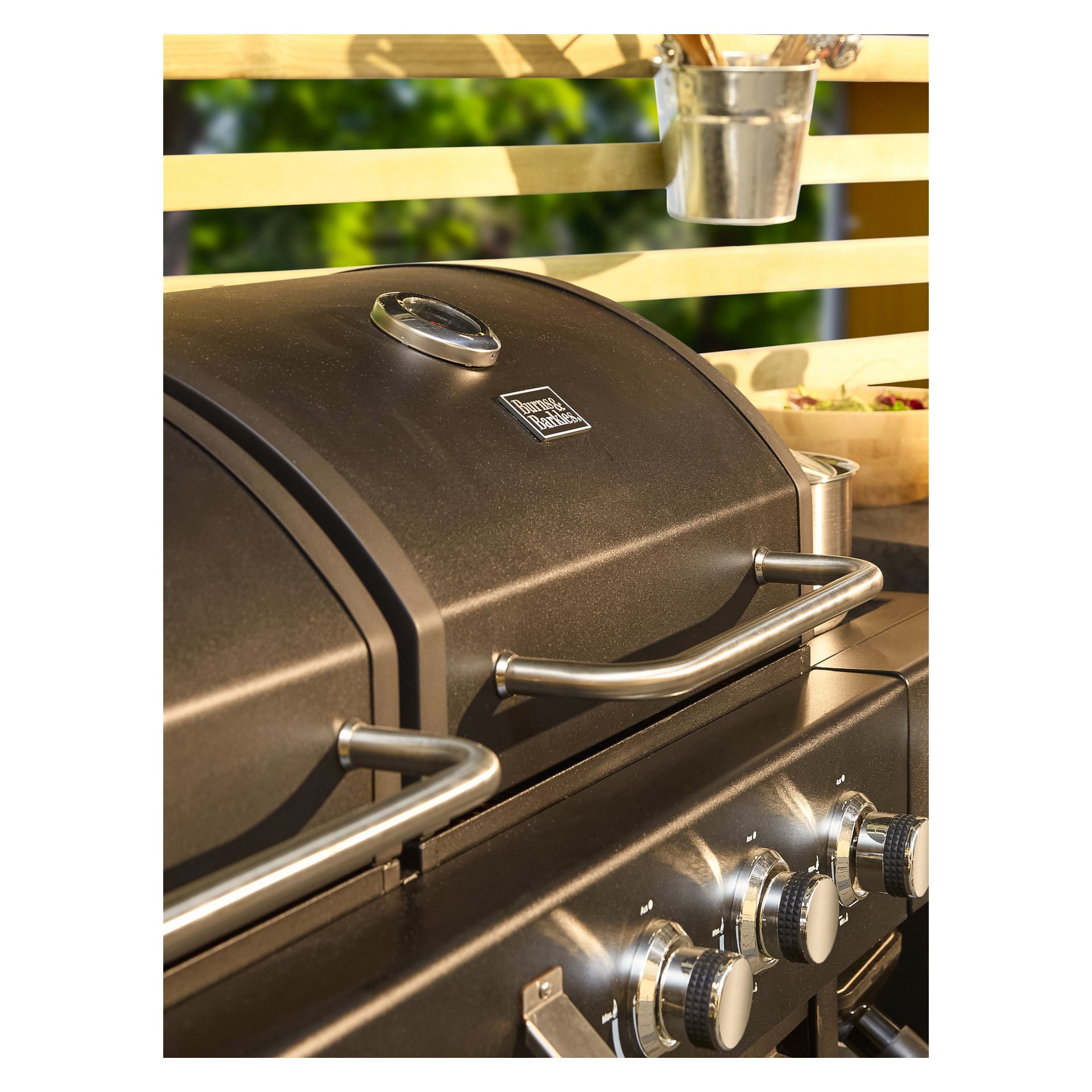 Burns & Barkles kol-/gasgrill All in One 8837 fångas i närbild och visar sin släta svarta yta, integrerade termometer, starka metallhandtag och kontrollrattar. Solljus lyser på grillens yta medan träribbor och en metallbehållare fylld med redskap står i bakgrunden.