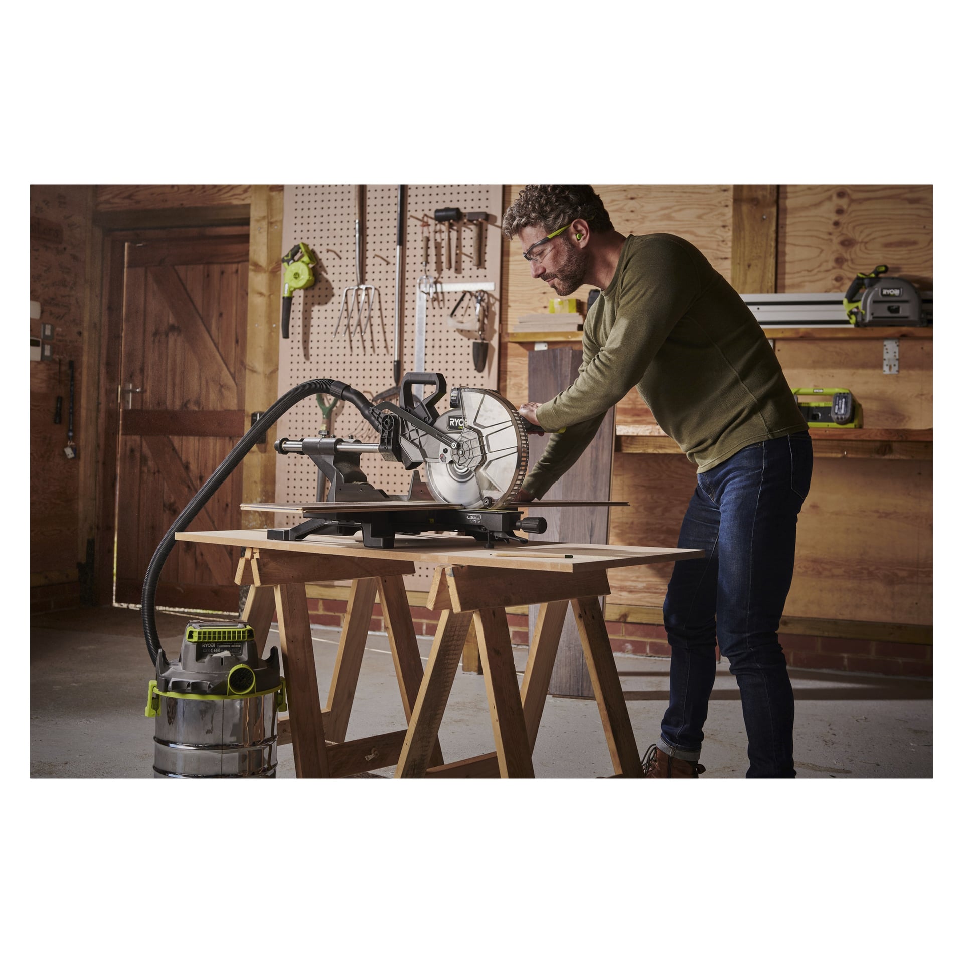 En man med skyddsglasögon använder en Ryobi One+ HP Mitre-såg RMS18254X-0 på en träarbetsbänk inne i en verkstad, med verktyg hängande på väggen och en verkstadsdammsugare bredvid bordet.