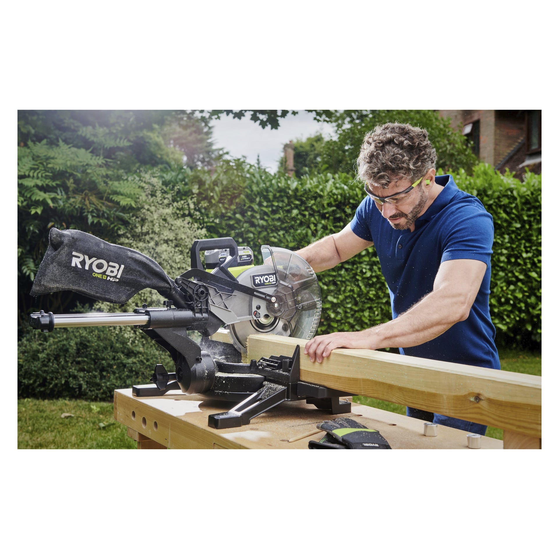 En person utrustad med skyddsglasögon och hörselskydd använder Ryobi One+ HP mitresåg RMS18254X-0 för att såga en träbalk utomhus. Det finns extra handskar på arbetsbänken, med träd och buskar synliga bakom dem.