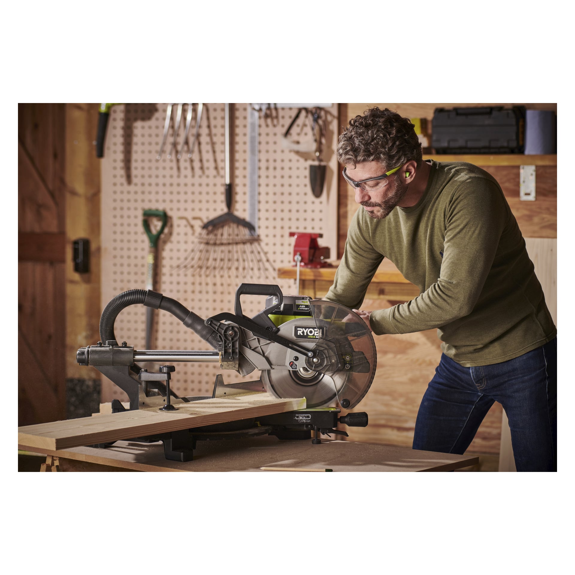 En man utrustad med skyddsglasögon och hörselskydd använder en Ryobi One+ HP mitresåg RMS18254X-0 för att såga trä i en verkstad, medan olika verktyg hänger på en pegboard bakom honom.