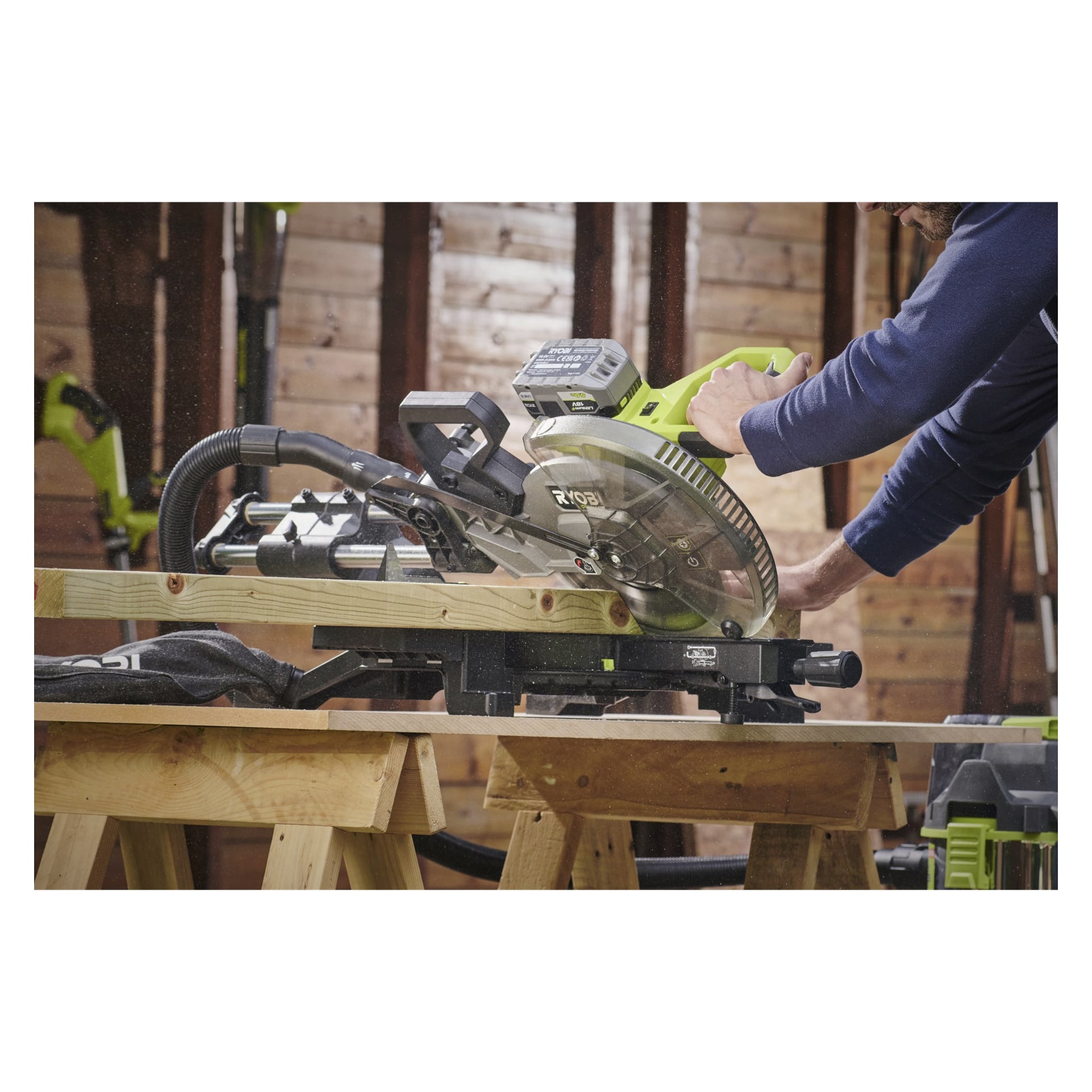 En person använder en Ryobi One+ HP mitresåg RMS18254X-0 för att såga en träplanka i en verkstad med träväggar. Sågspån är synligt i luften, och sågen vilar på en arbetsbänk.