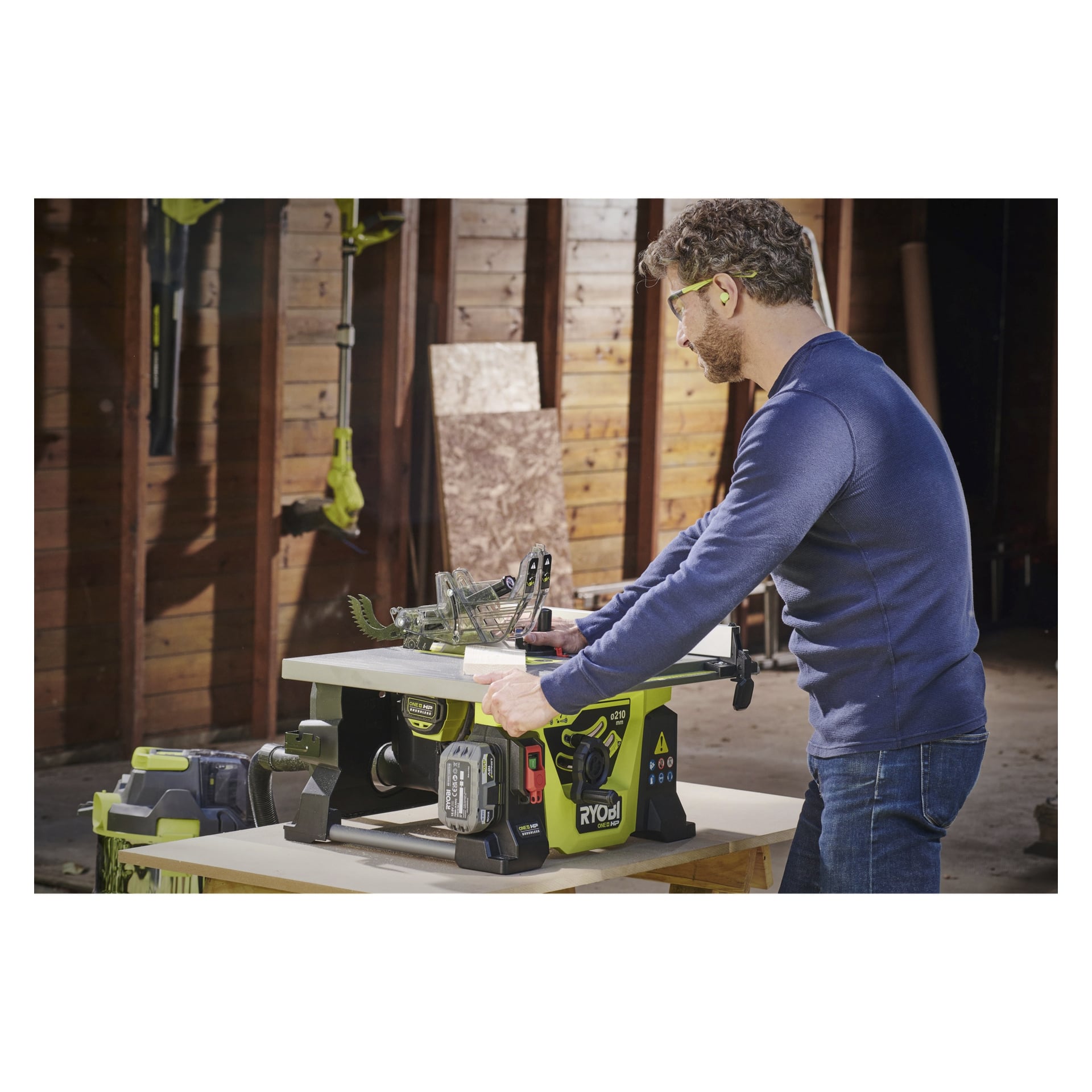 En man med skyddsglasögon och öronproppar använder en Ryobi One+ HP batteridriven bordsåg modell RTBS18X-0, och styr en träskiva genom bladet i en verkstad av trä. Han står nära sågen medan han gör snittet.