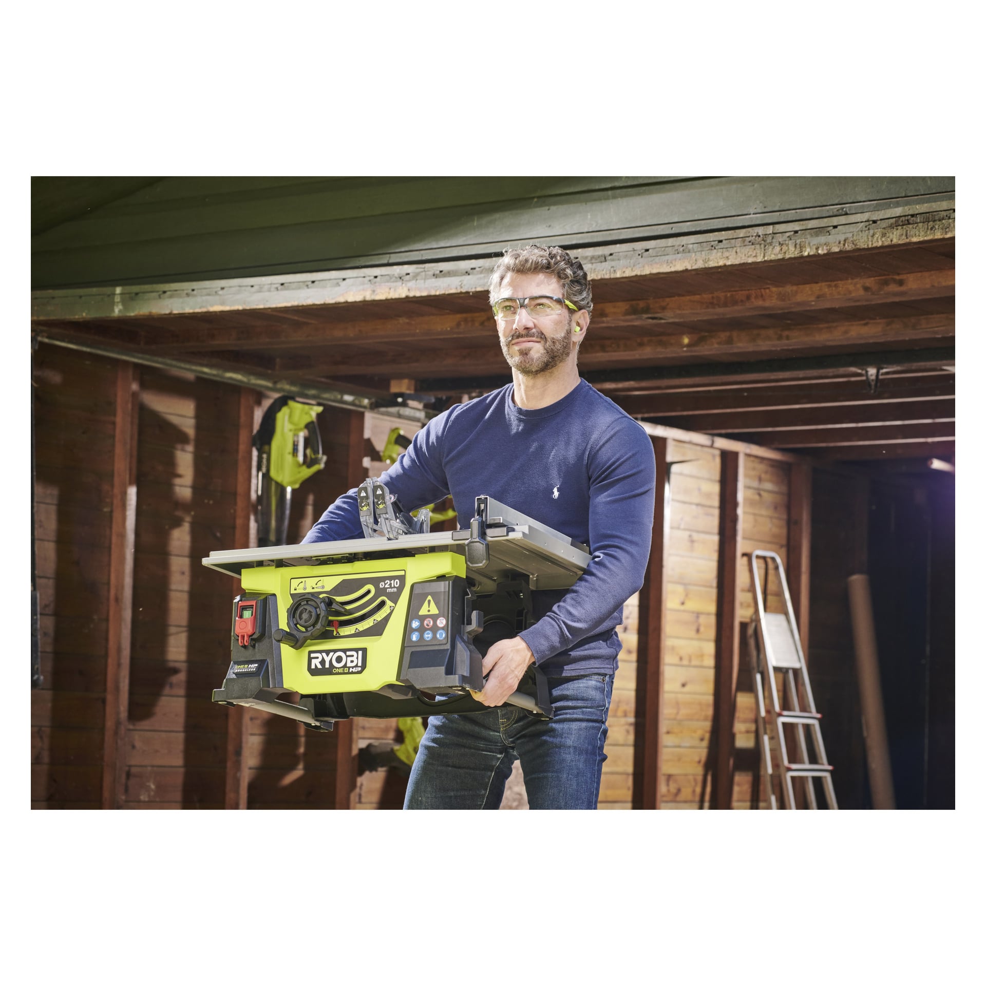 En man med skyddsglasögon och en blå långärmad skjorta håller en grön Ryobi One+ HP batteridriven bordsåg RTBS18X-0 inne i en träverkstad, med olika verktyg och en stege i bakgrunden.