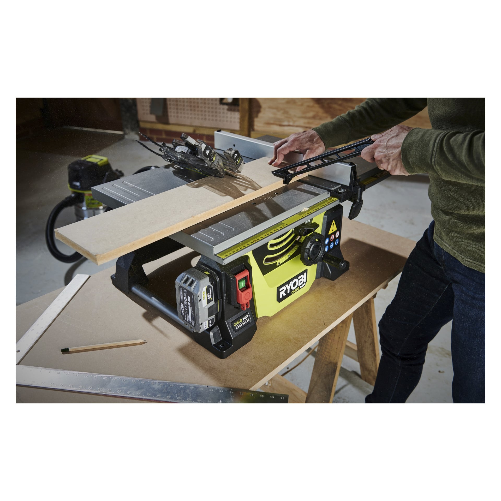 Någon använder en Ryobi One+ HP batteridriven bordsåg RTBS18X-0 för att såga trä i en verkstad, där sågen vilar på en arbetsbänk omgiven av olika verktyg och material.