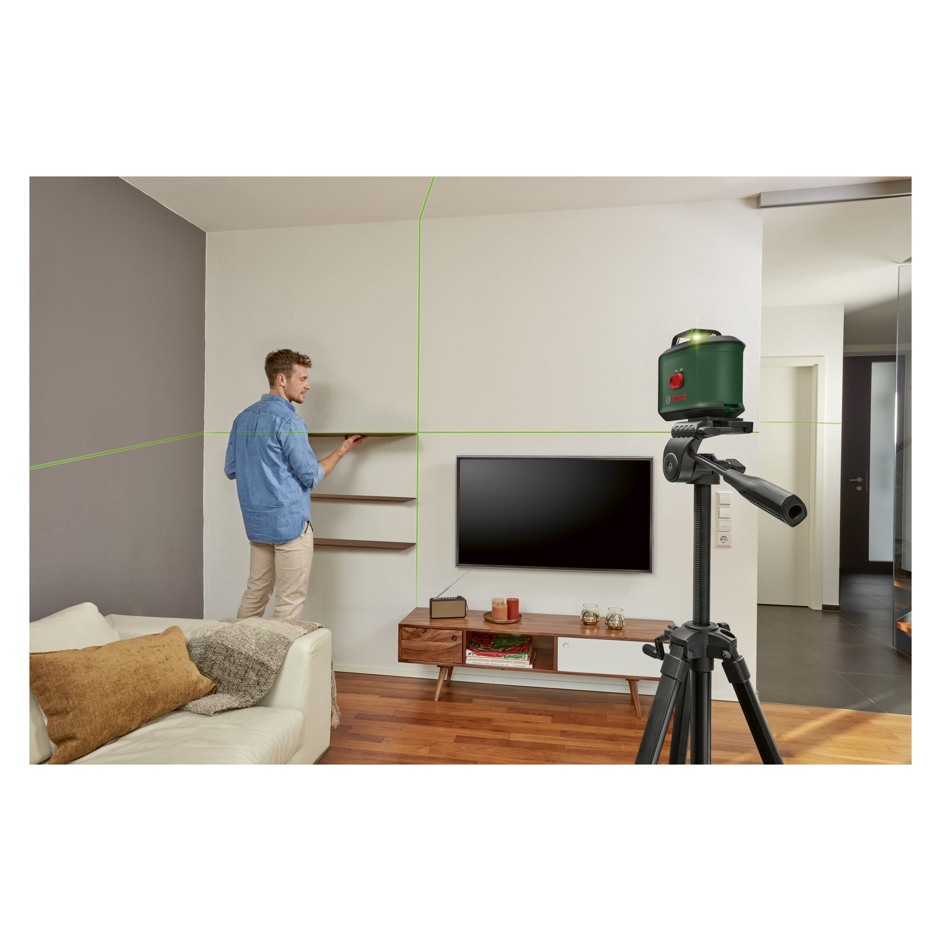 En man placerar hyllor ovanför en TV i ett modernt vardagsrum och använder Bosch Cross-lasern UniversalLevel 360 monterad på ett stativ. Rummet har trägolv och moderna möbler.