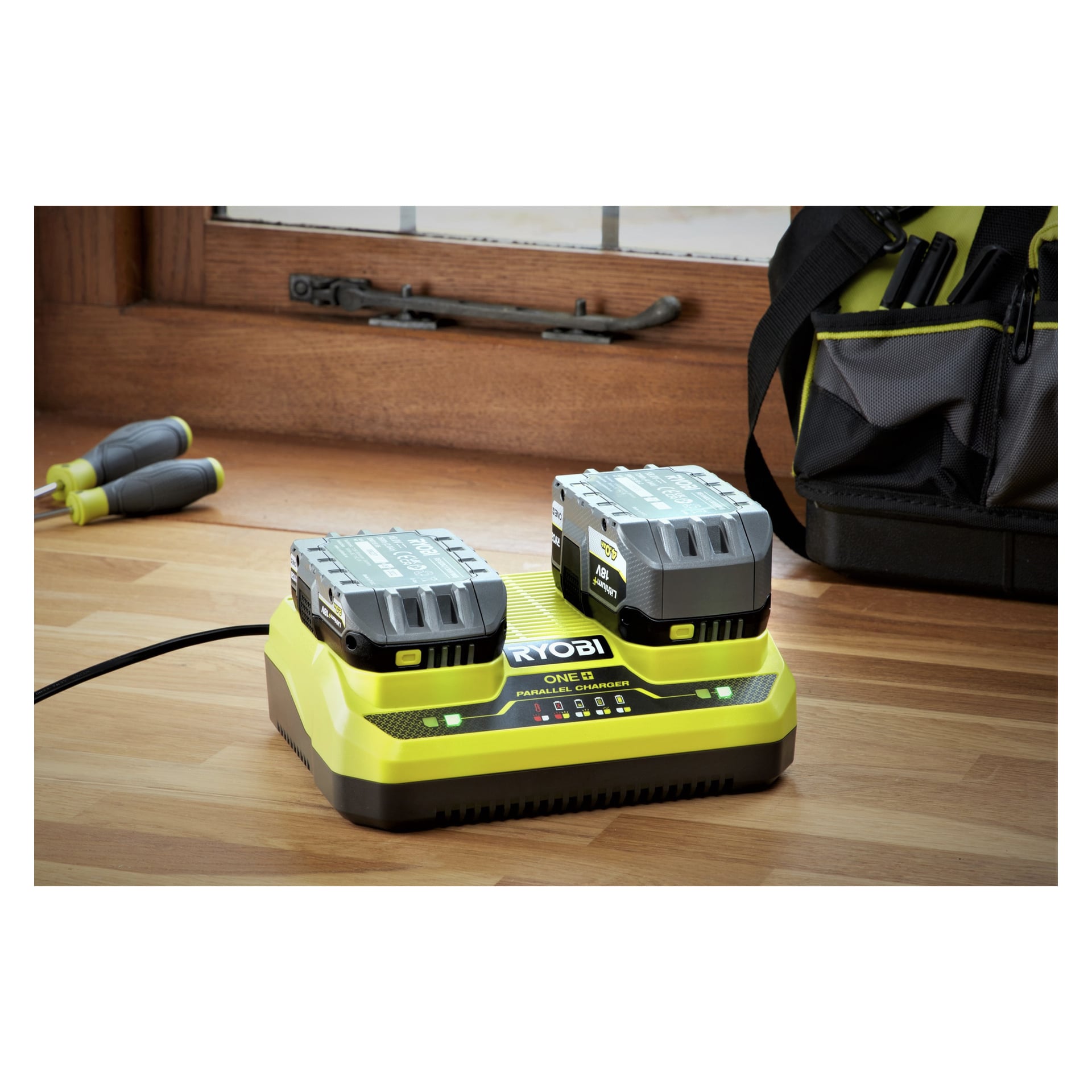 En Ryobi One+ Double-laddare håller två batterier som laddas på ett trägolv vid ett fönster, med två skruvmejslar och en verktygsväska i bakgrunden.