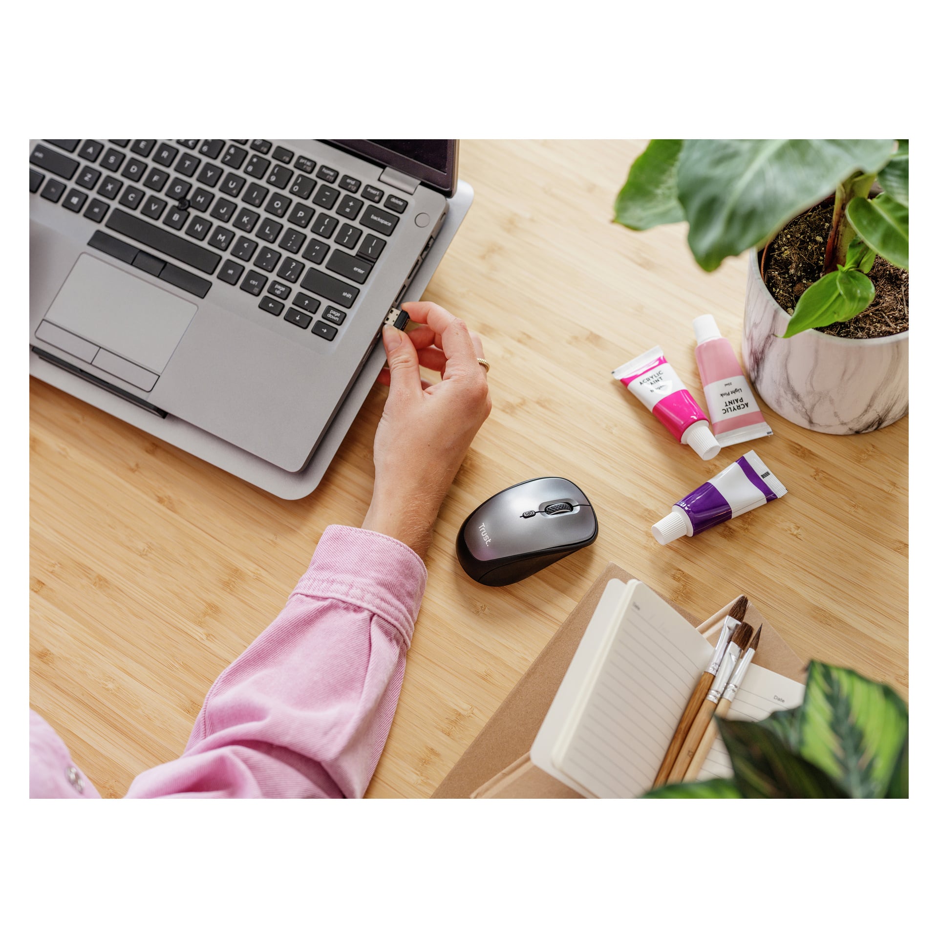 Iklädd en rosa skjorta kopplar någon in en USB-enhet i en laptop på ett träbord. En Trust trådlös mus, några färgtuber, en anteckningsbok med pennor och en krukväxt står i närheten.