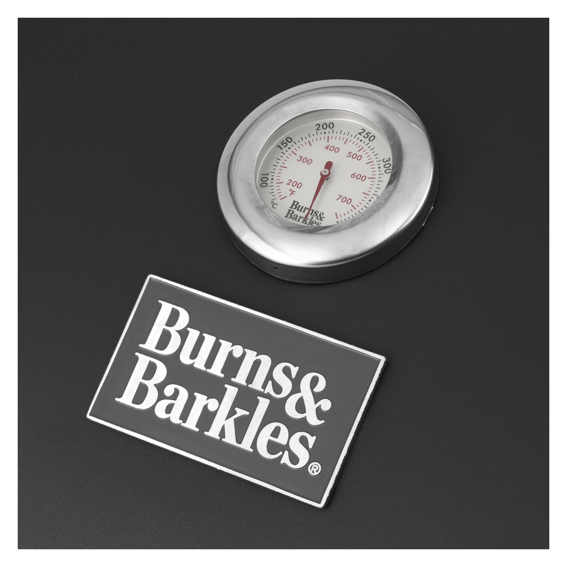 En cirkulär metalltermometer som visar både Fahrenheit och Celsius är placerad på en mörk yta bredvid en rektangulär Burns & Barkles-skylt, bredvid en Charcoal Grill Coal King 5641 från samma märke.