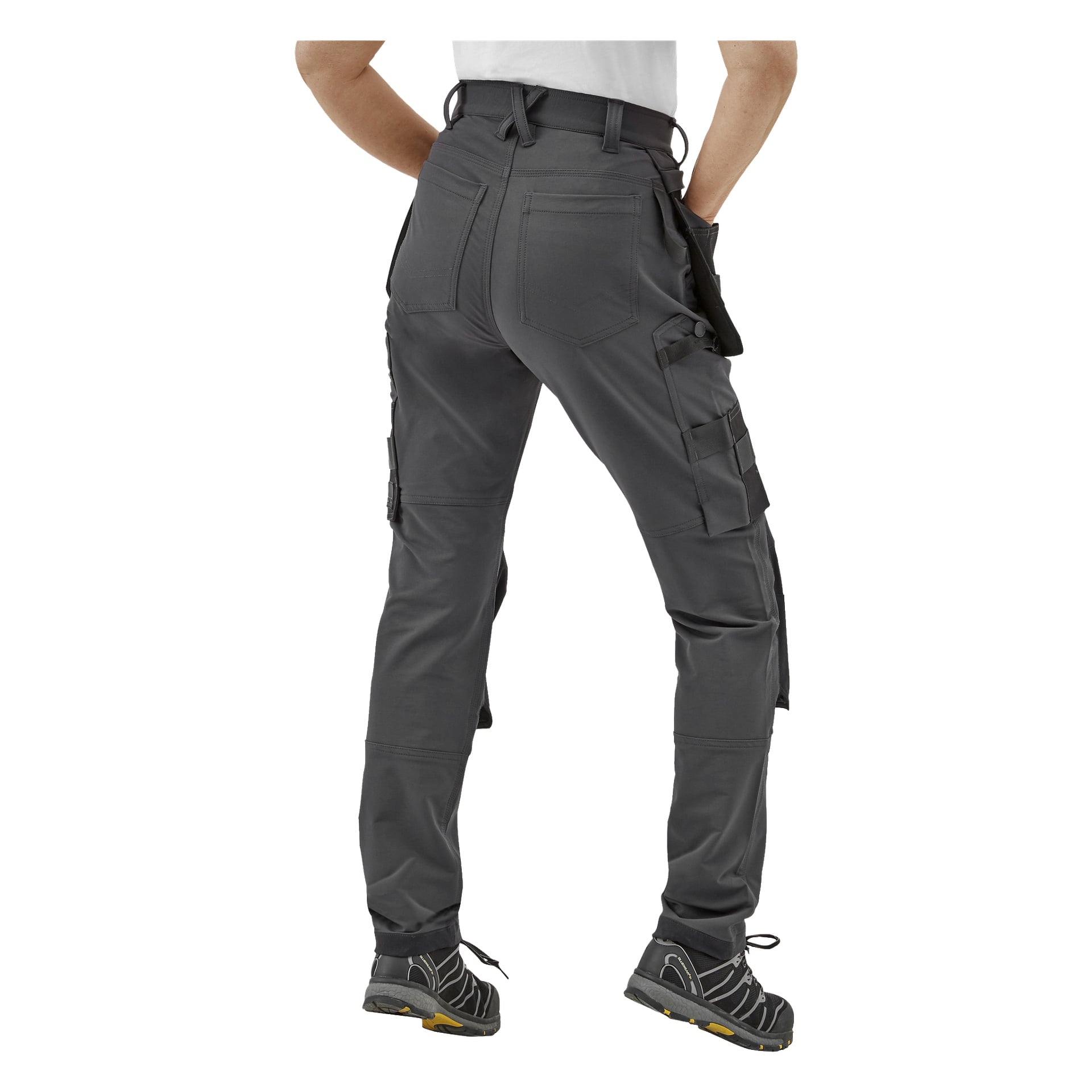 En person står med ryggen mot kameran, iklädd en vit skjorta och mörkgrå Bluewear Pro Workwear Dambyxor Stella med flera fickor och förstärkta knän, tillsammans med svarta och gula skor.