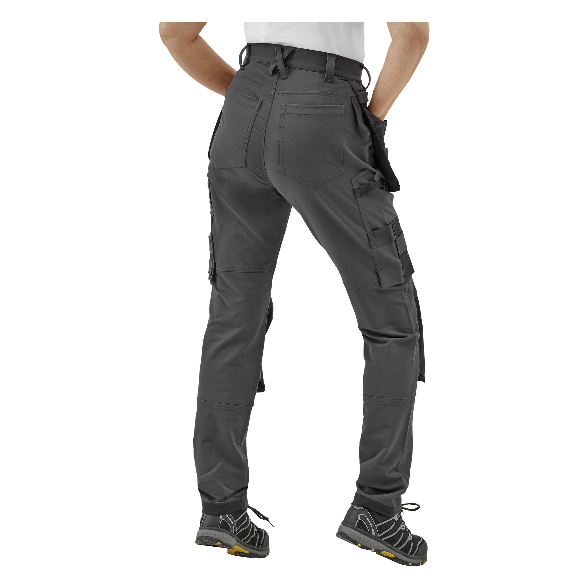 En person syns bakifrån, stående med benen lätt isär, iklädd Bluewear Pro Workwear Women's work pants Stella i mörkgrått med flera fickor och öglor, svarta och gula arbetsskor samt en instoppad vit skjorta.