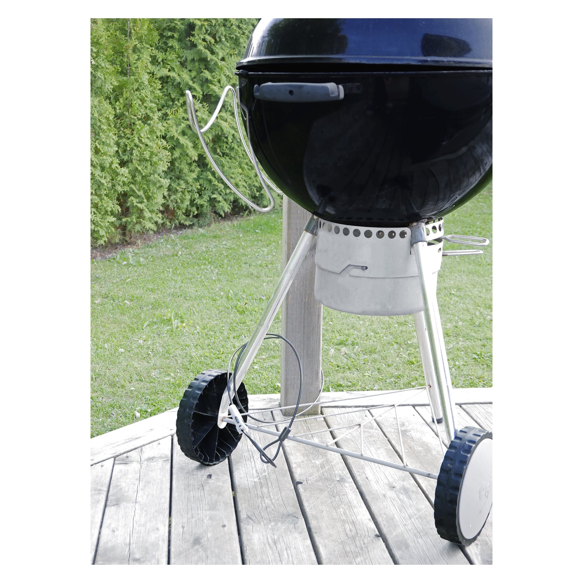 Ett ABUS Combiloop trådlås säkrar ett svart kolgaller med lock på toppen, placerat på ett trädäck. Grillen har två framträdande hjul och stadiga metallben. Bakom grillen finns en grön gräsmatta omgiven av träd.