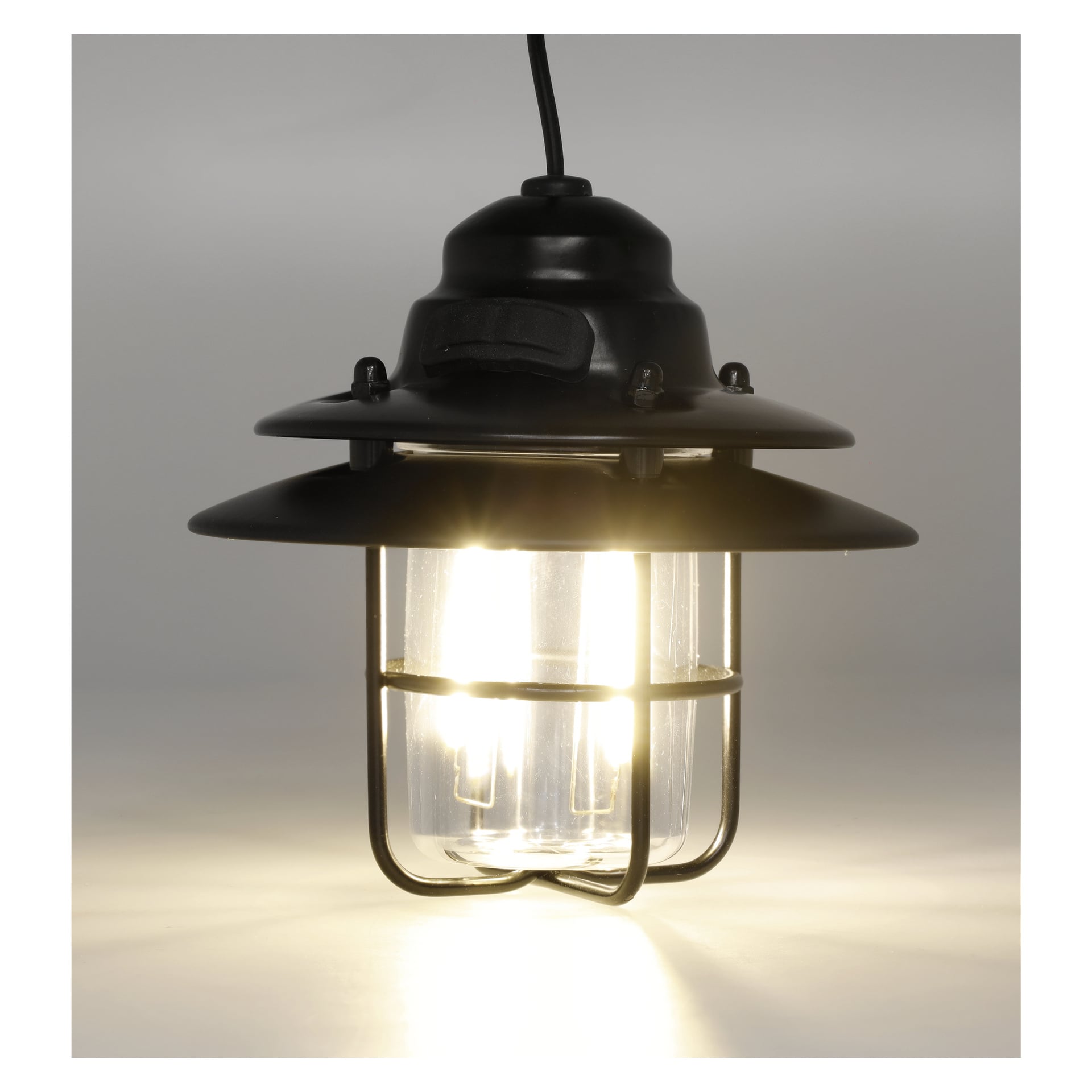 En Kayoba Rechargeable campinglampa med svart metallkropp och glasskärm, innesluten i en skyddande metallbur, hänger tänd med en svart sladd.