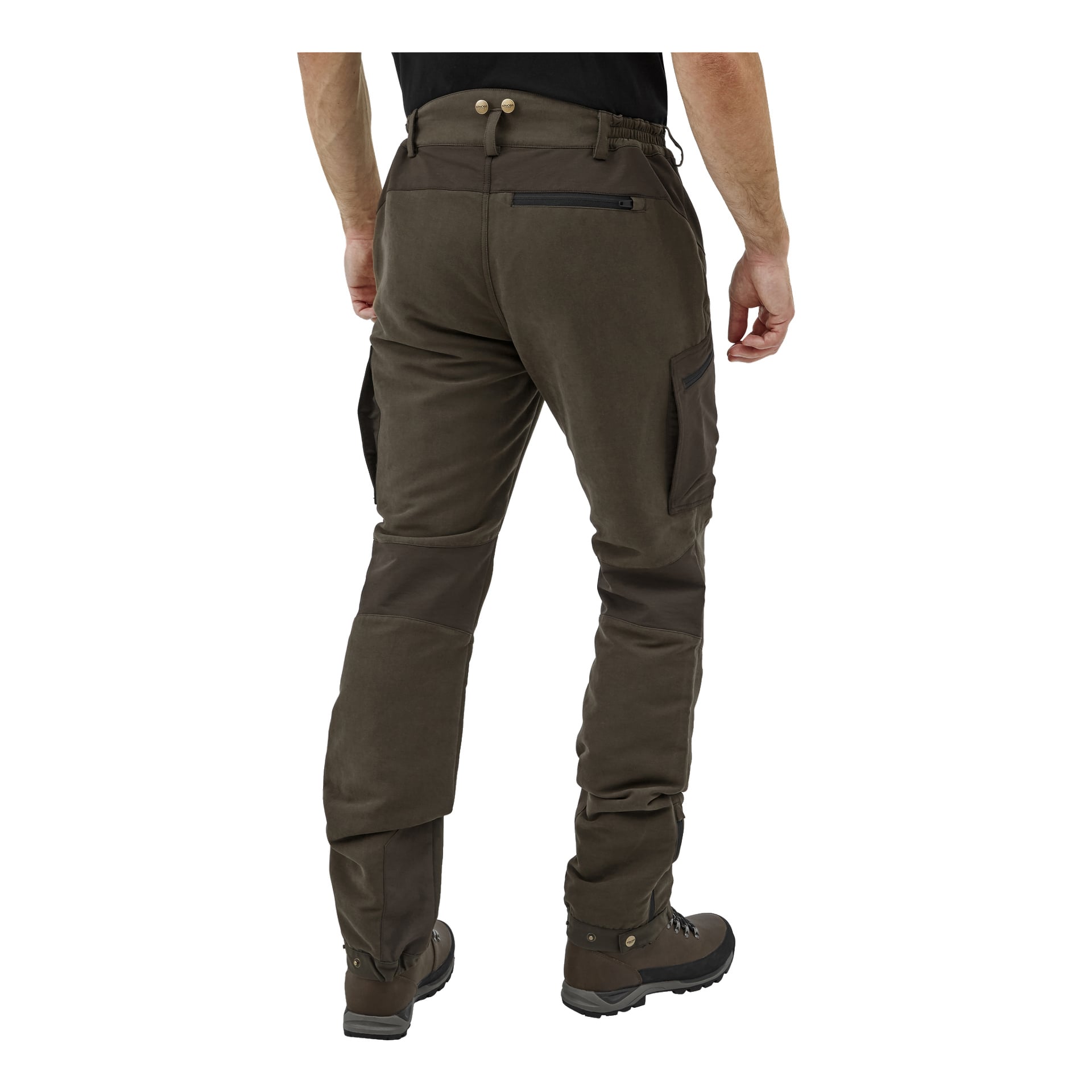 En person står med ryggen mot kameran, klädd i bruna Kayoba Outdoor Men's Grimsö jaktbyxor med flera fickor och bruna vandringskängor. Byxorna har förstärkta sektioner på knäna och sätet, samt en ficka med dragkedja på vänster sida.