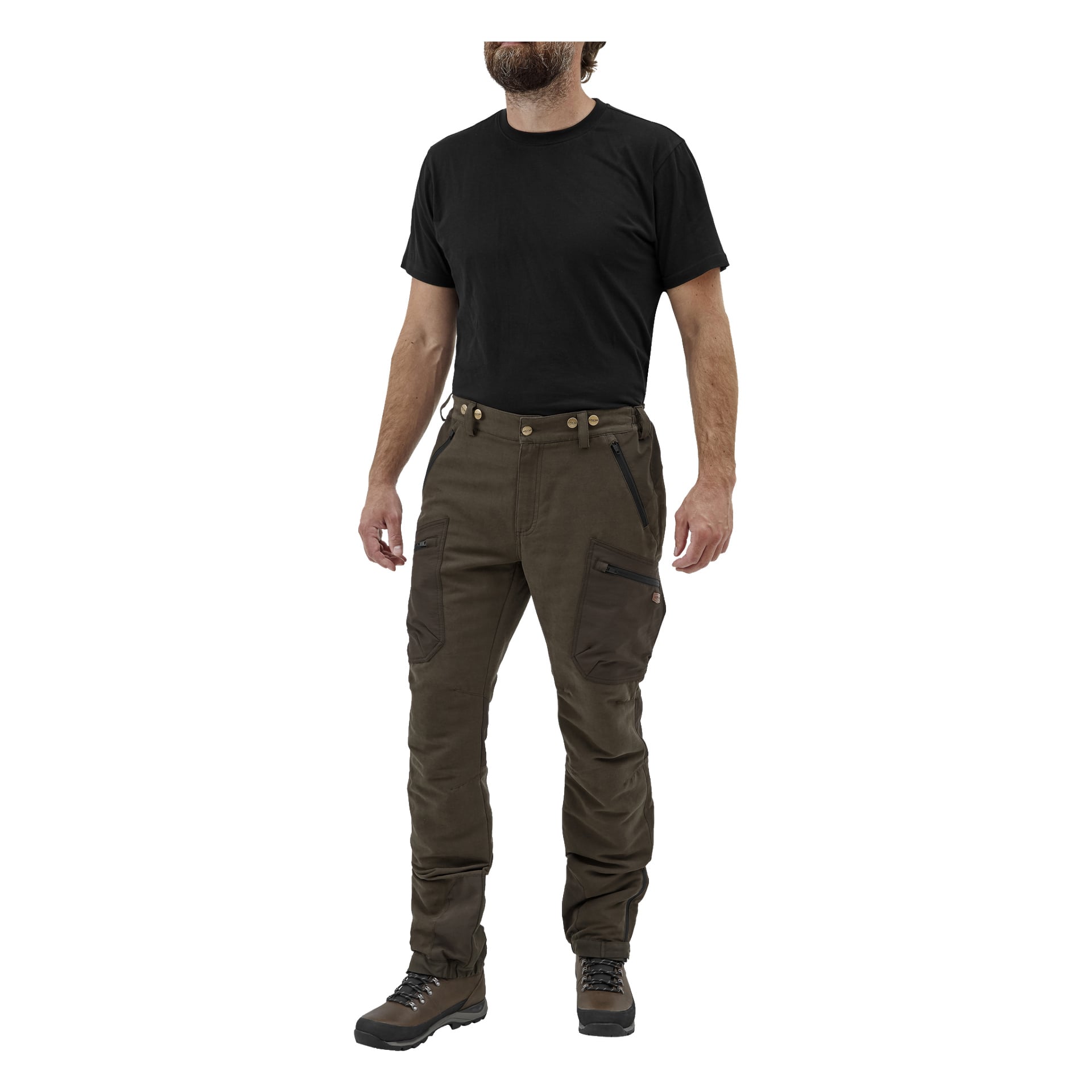 En man står något åt vänster, klädd i en enkel svart t-shirt, Kayoba Outdoor Men's Grimsö jaktbyxor med lårfickor och dragkedjor, samt bruna vandringskängor. Hans armar vilar naturligt vid sidorna och ansiktet syns bara delvis.