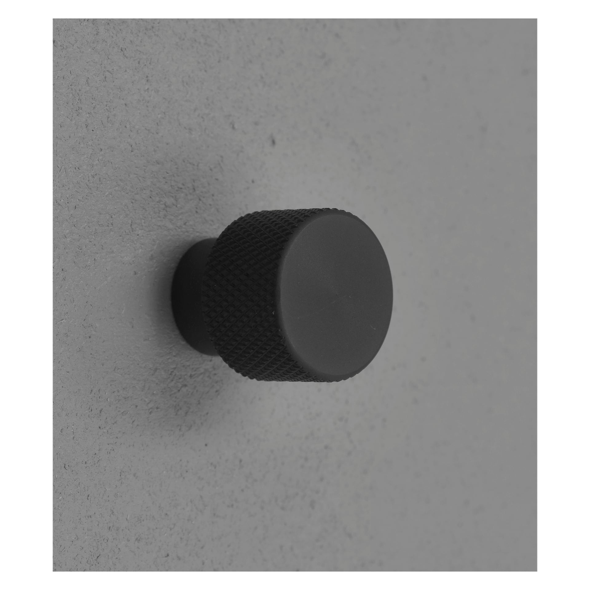 En svart, rund Hard Head Button fra Knob 2-pakken er festet til en lys grå vegg. Den riflede teksturen forbedrer grepet, og bildet viser den i en svak vinkel.