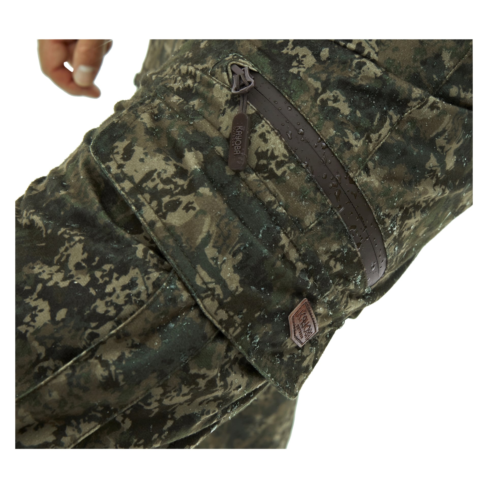Detaljerad vy av Kayoba Outdoor Men's Cannock Superior jaktbyxor, med framhävning av kamouflagedesignen, en brun dragkedjasficka och en liten brun tyglapp med synliga vattendroppar. Övre vänstra delen visar en del av bärarens hand.