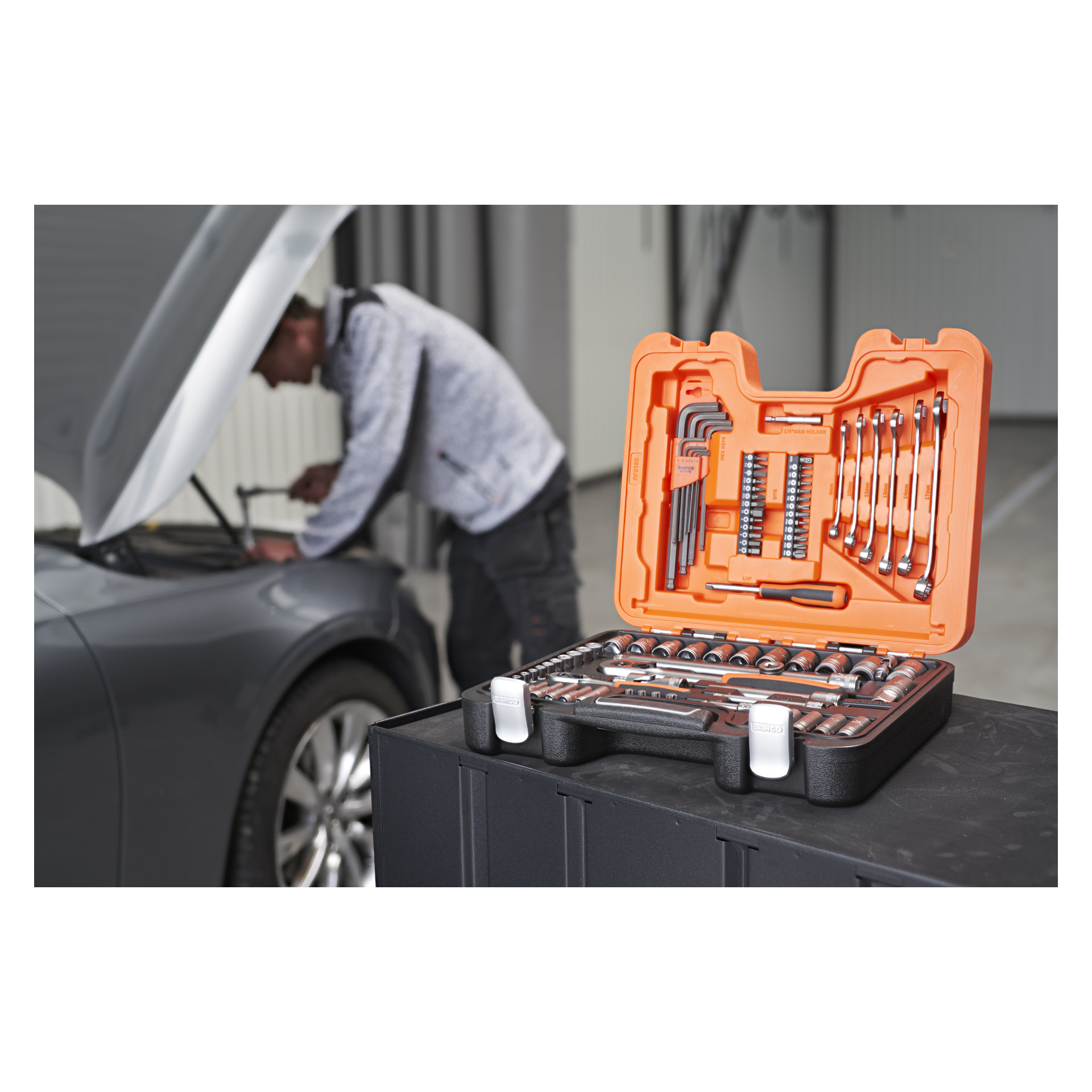 Bahco Socket Wrench Set S910-JU, jossa on 91 kappaletta, on esillä avoimena musta-oranssissa kotelossa pinnalla. Sen takana joku työskentelee harmaan auton konepellin alla autotallissa tai työpajassa.