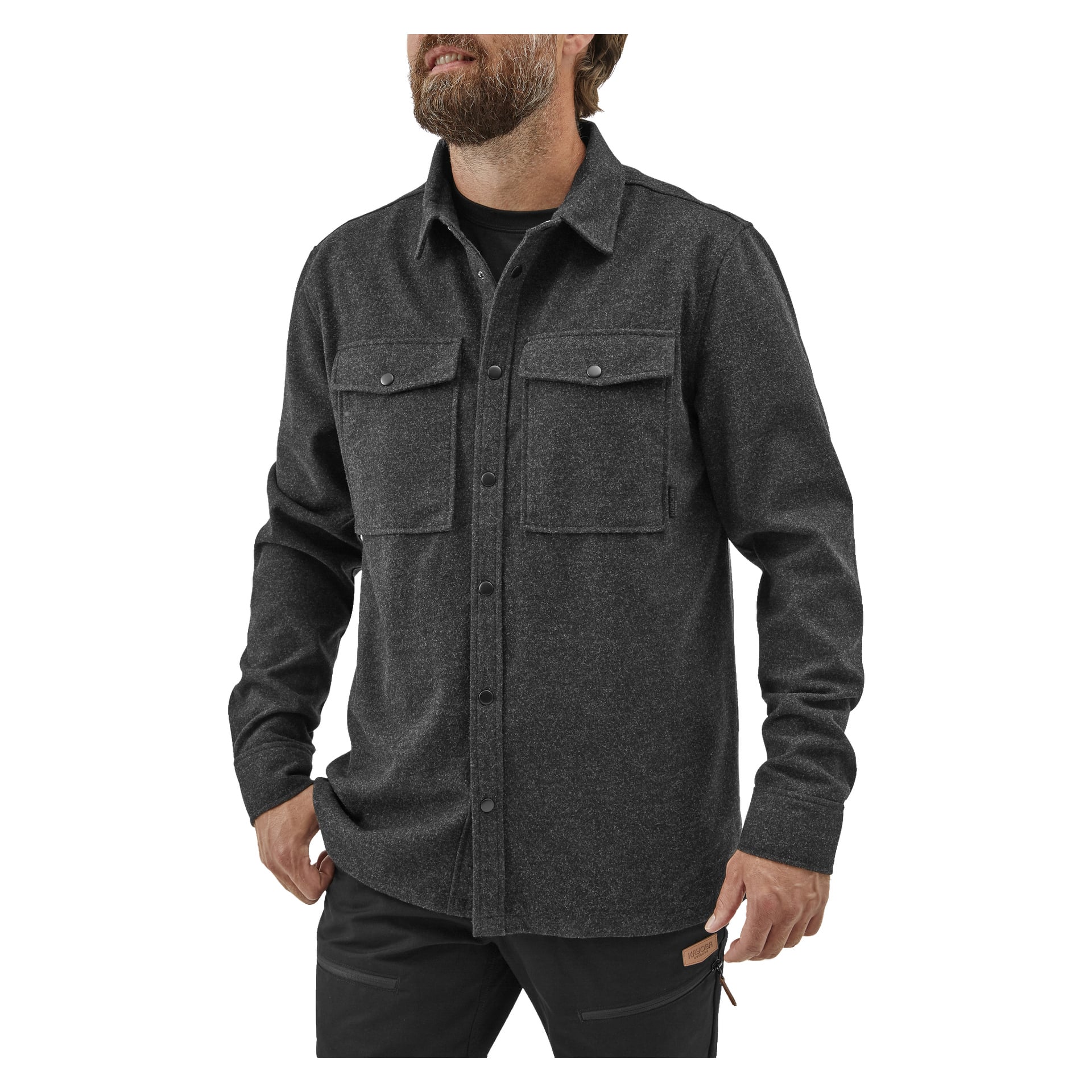 Parrakas mies, jolla on lyhyet hiukset, seisoo toinen käsi taskussa, pukeutuneena tummanharmaaseen Kayoba Outdoor Overshirt -miesten Idhultiin, mustaan aluspaidan ja mustiin housuihin pukeutuneena. Hänen kasvonsa näkyvät osittain.