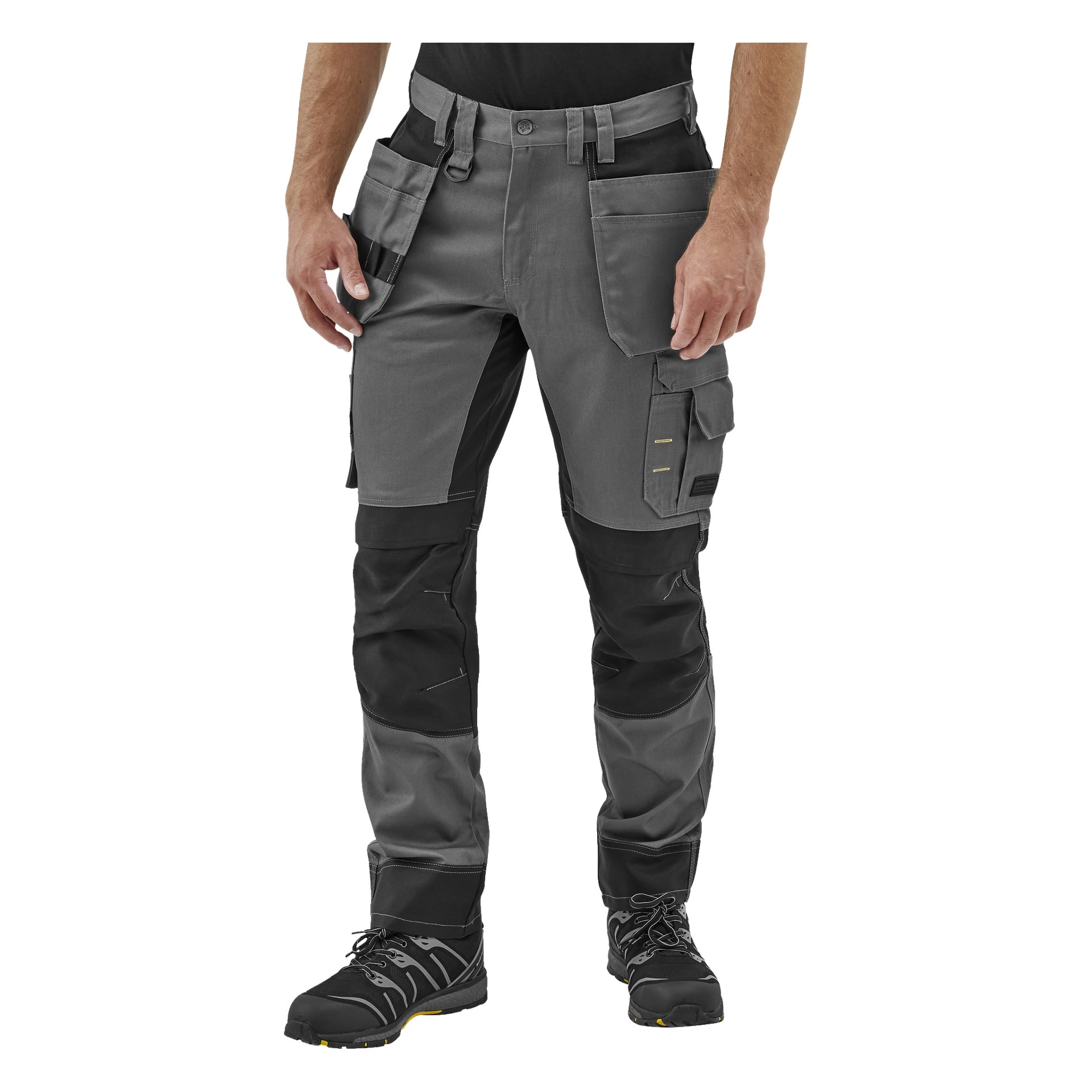 En vy från midjan och ner av någon som bär grå och svarta Bluewear Rex arbetsbyxor för herrar med förstärkta knän och flera fickor, tillsammans med svarta arbetsskor.
