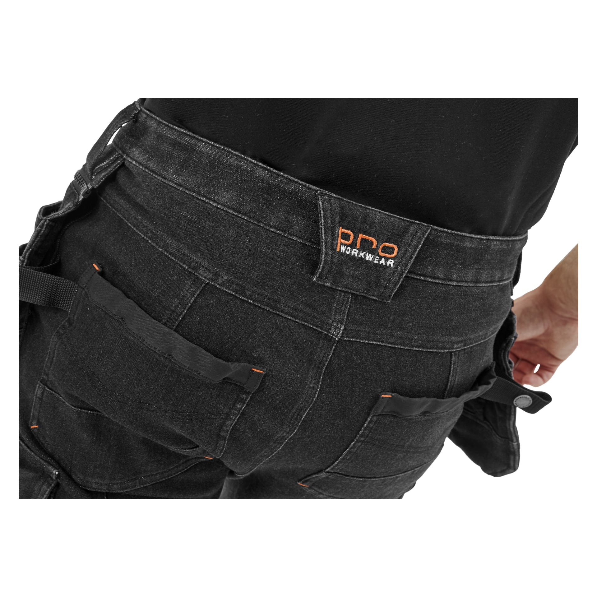 Närbild på en person i Bluewear Pro Workwear herrbyxor Steve Denim. De svarta byxorna har flera fickor, markerade med orange sömmar, och visar en midjebandsetikett som säger PRO WORKWEAR. Personens hand verkar delvis synlig vid höften.