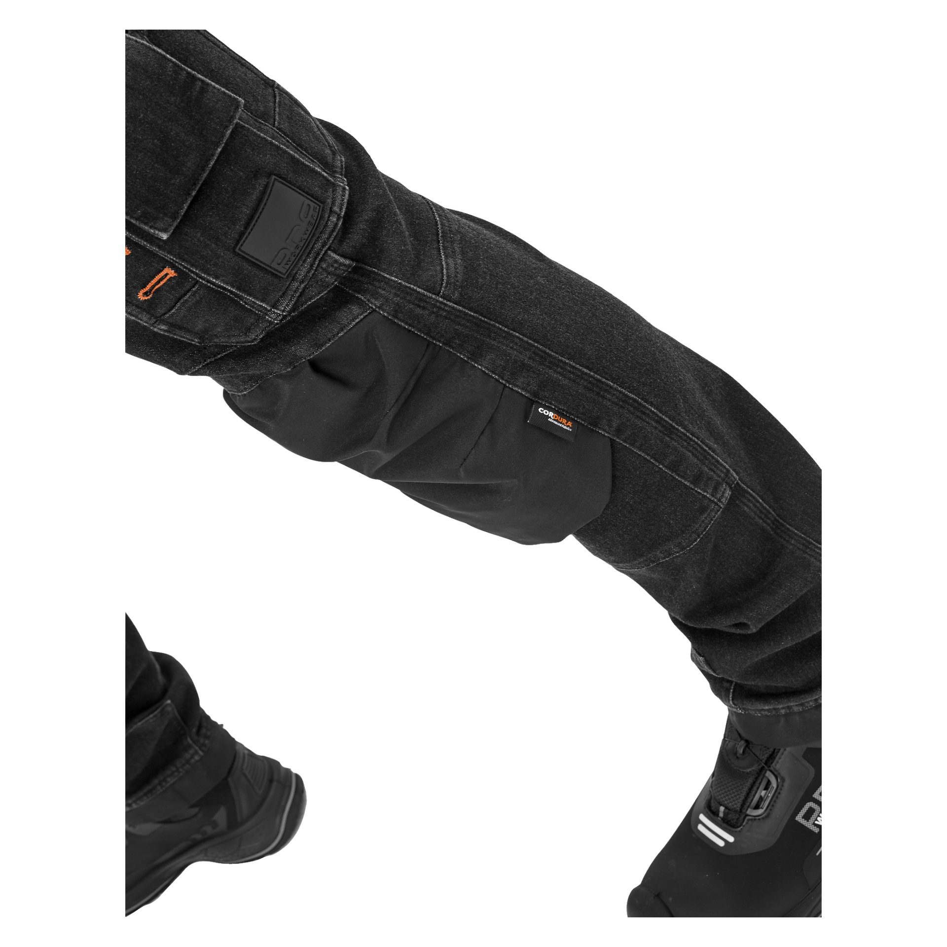 En närbild på en persons böjda ben klädd i Bluewear Pro Workwear Steve Denim-byxor för män, med förstärkta knäskydd, detaljerade synliga stygn, en orange etikett på knät och bärs med svarta motorcykelstövlar.