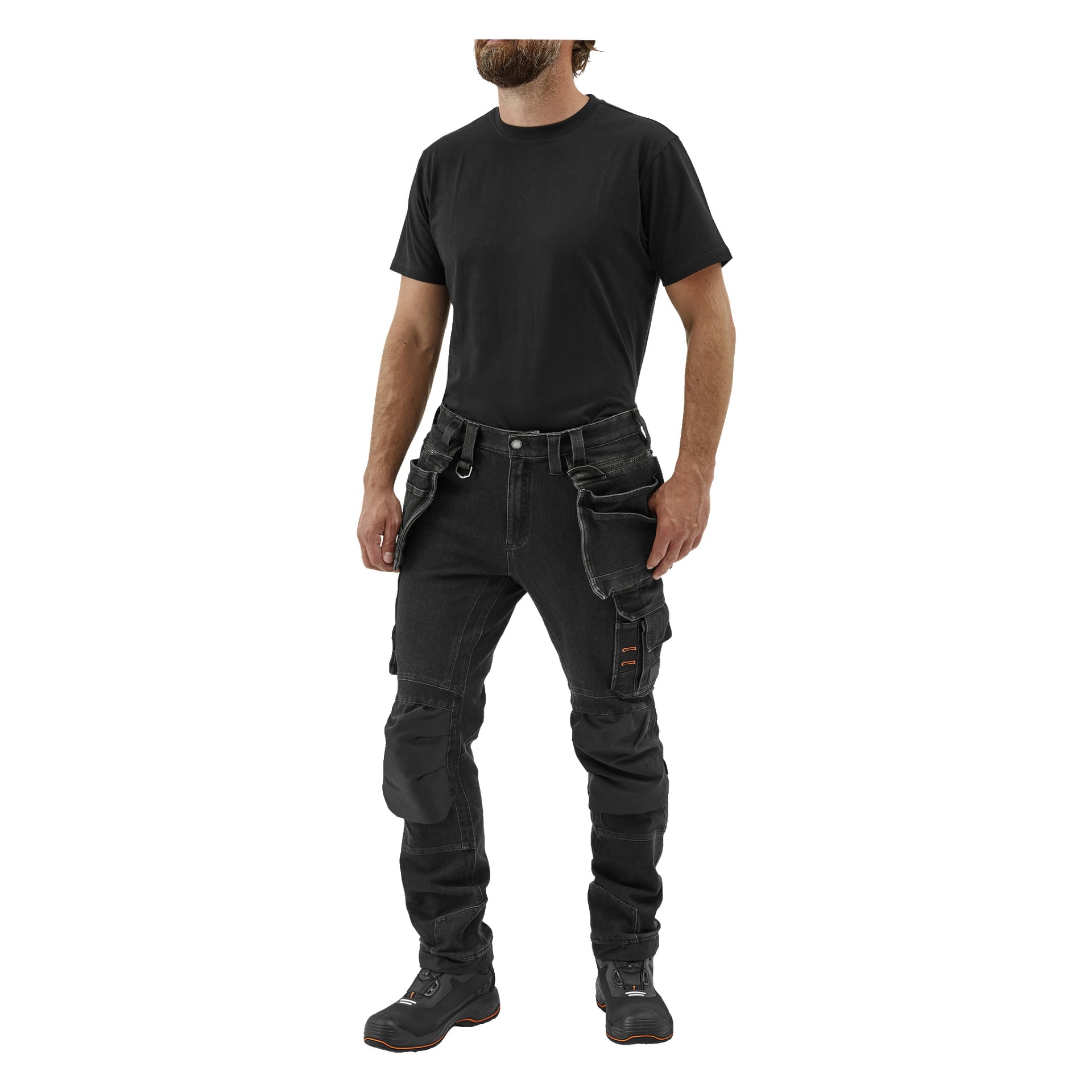 En man står klädd i en svart t-shirt, Steve Denim Bluewear Pro Workwear herrbyxor med flera fickor och knäskydd, tillsammans med svarta arbetsskor. Hans armar hänger längs sidorna och huvudet är lätt vridet.