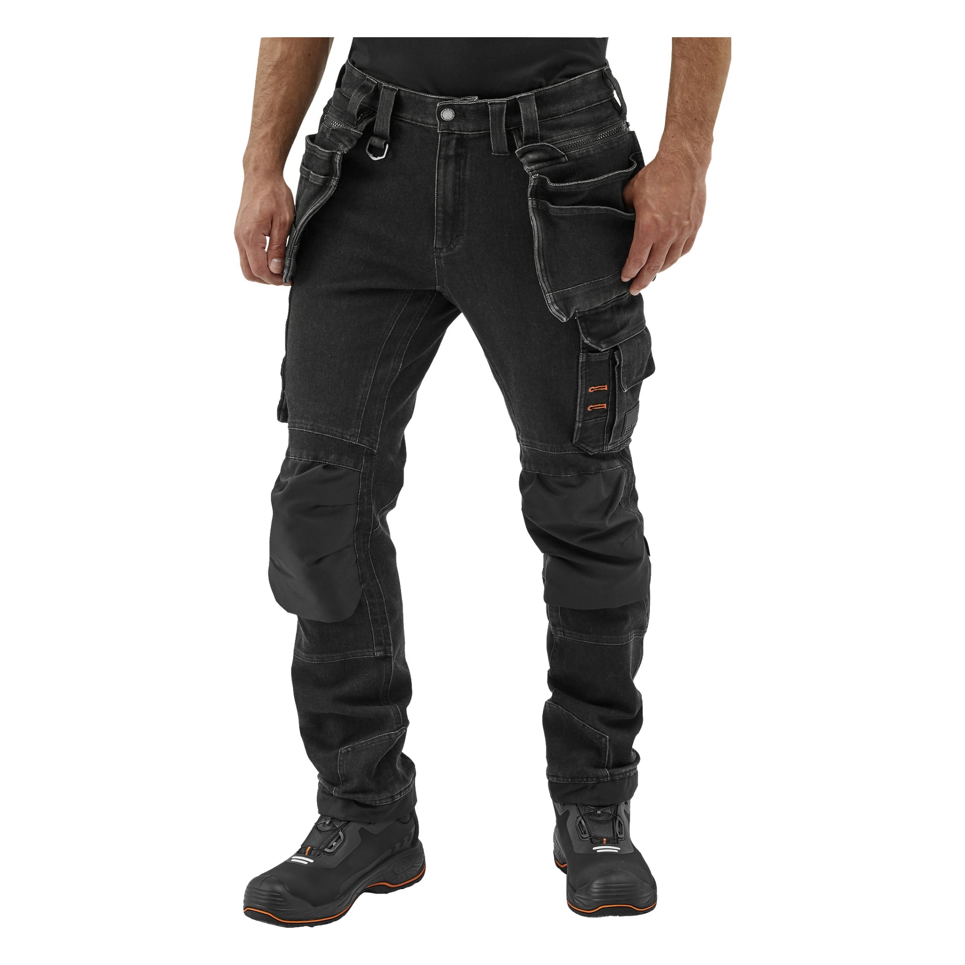En person visas från midjan och neråt, klädd i Blackwear Blue Pro Workwear Steve Denim-byxor med flera fickor och förstärkta knän, tillsammans med svarta arbetskängor.