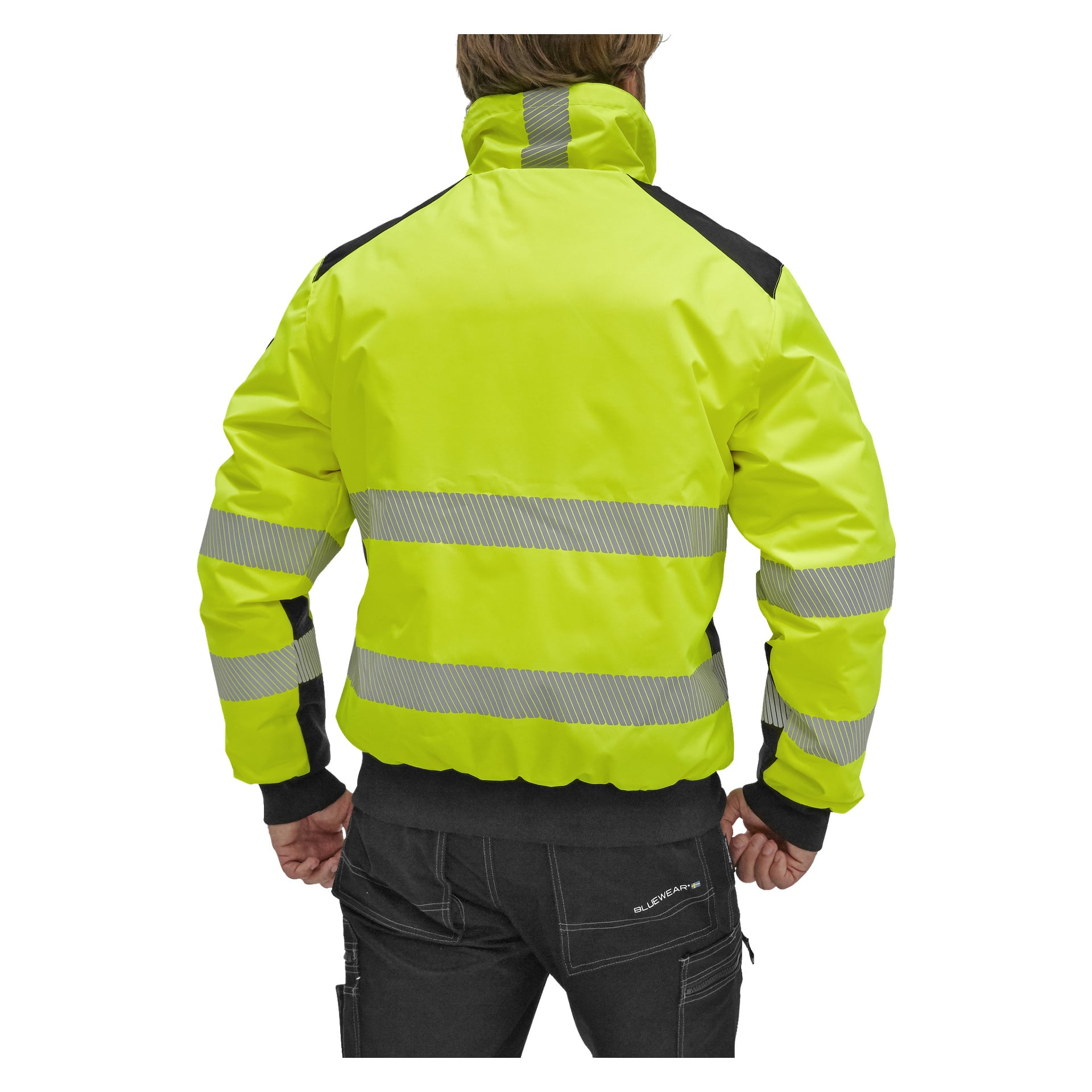 Någon står med ryggen mot kameran, klädd i en Bluewear Warm-fodrad Hi-Vis Hamilton-jacka. Jackan är klargul med reflekterande horisontella ränder och svarta detaljer, tillsammans med mörka byxor.