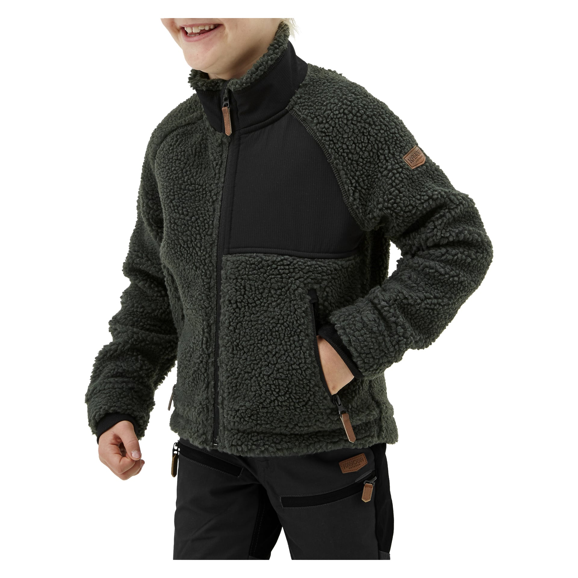 Ett leende barn bär Kayoba Outdoor Children's pilejacka gjord av mjuk, texturerad mörkgrön fleece med dragkedja fram, två fickor med dragkedja och svarta lappar på axlarna. Barnet har också mörka byxor.
