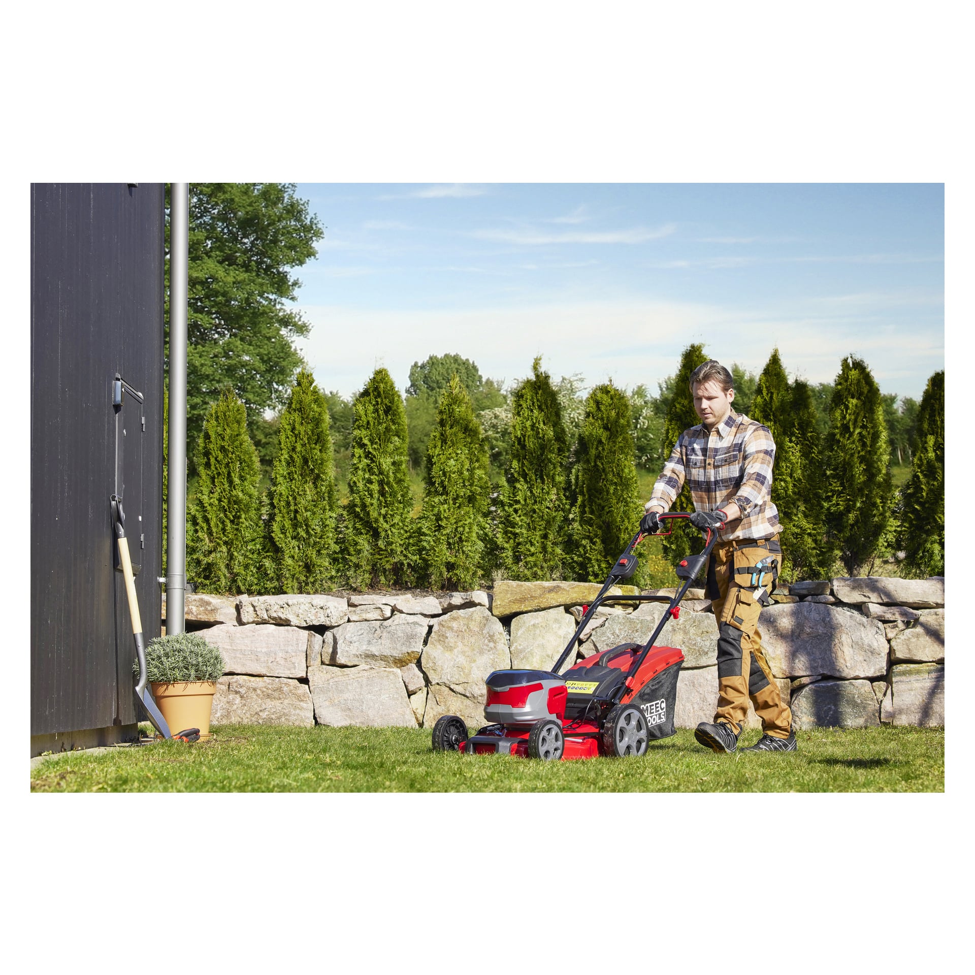 Osoba w koszuli w kratę i khaki spodniach kosi trawnik samobieżną kosiarką bateriową Meec Tools Multiseries 40 V przy kamiennym murze, podczas gdy łopata opiera się o ciemny budynek.