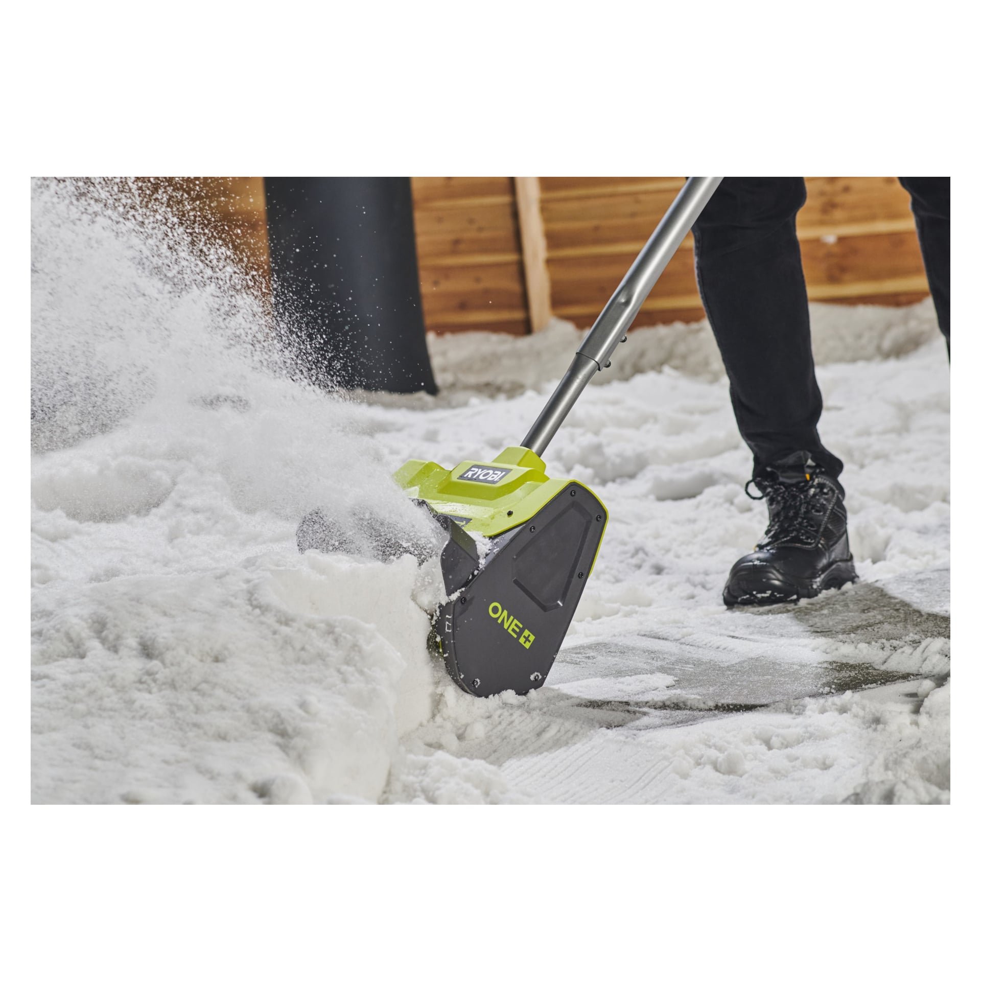En person som kör en Ryobi One+ RY18ST25A-0 batteridriven snöskyffel röjer bort snö när den kastas åt sidan. Personen är klädd i svarta byxor och stövlar, med en trävägg bakom sig.