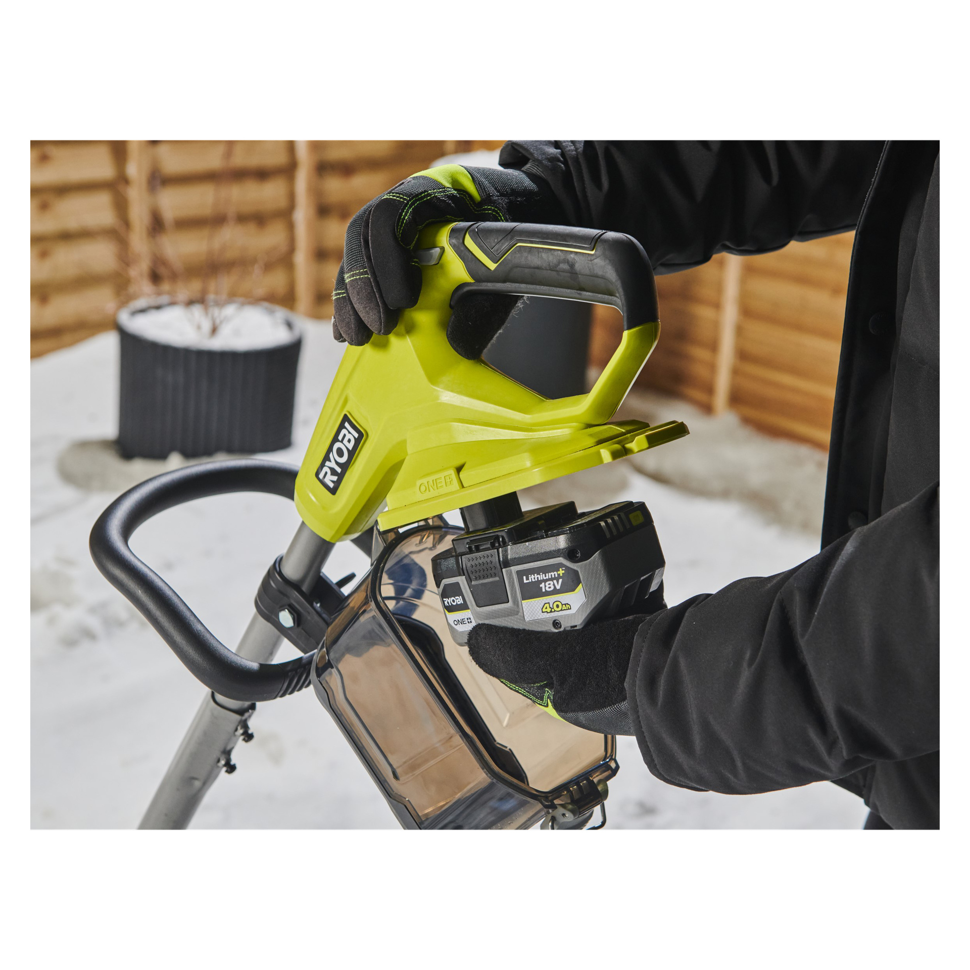 Med svarta handskar skjuter en person in ett 18V Ryobi One+ Lithium+ 4,0 Ah-batteri i ett gult Ryobi One+ RY18ST25A-0 elverktyg utomhus på snö, med ett trästaket bakom sig.