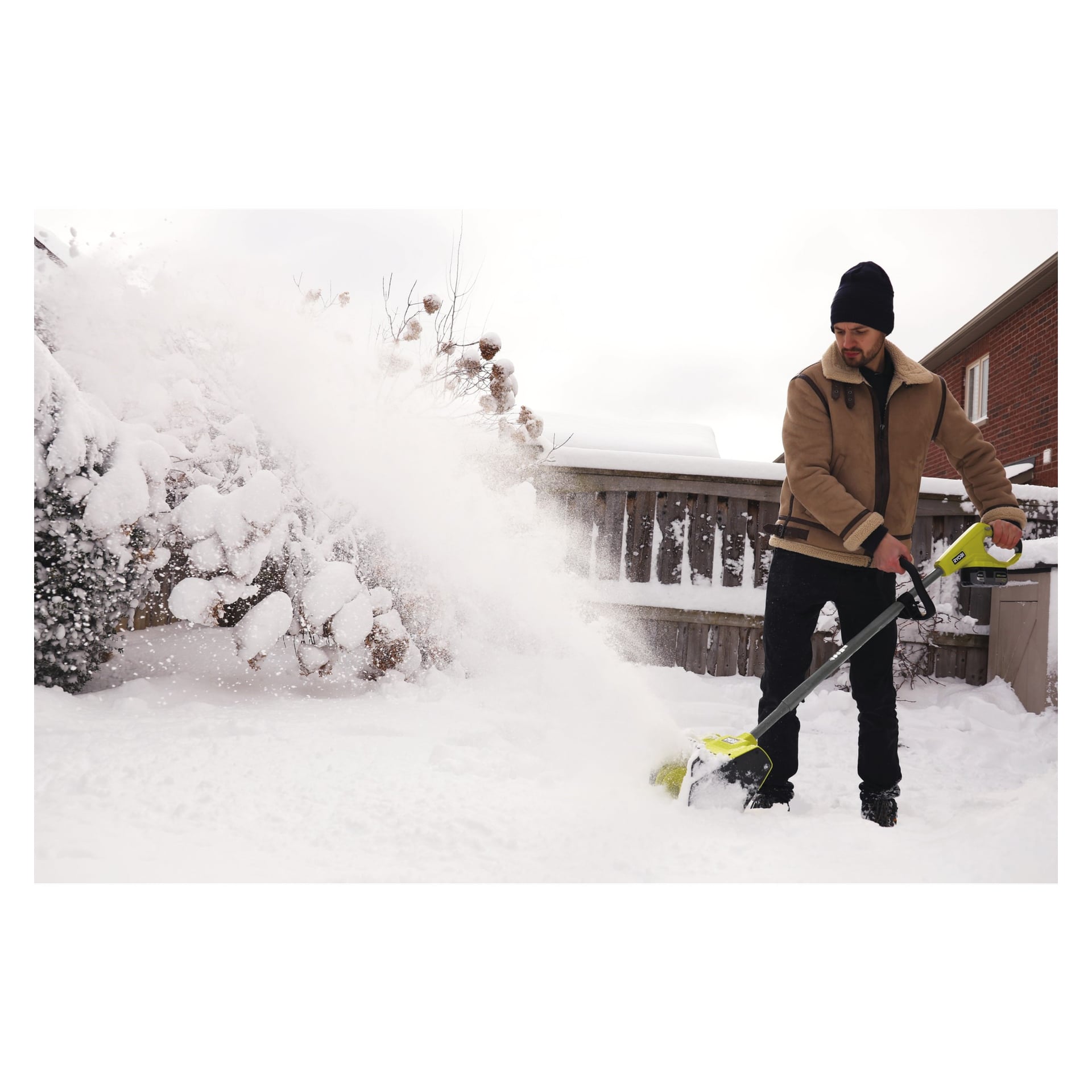 I vinterkläder använder man en grön Ryobi One+ RY18ST25A-0 snöslunga för att ta bort snö från en bakgård. Området omges av ett trästaket och buskar, med snö som täcker marken.