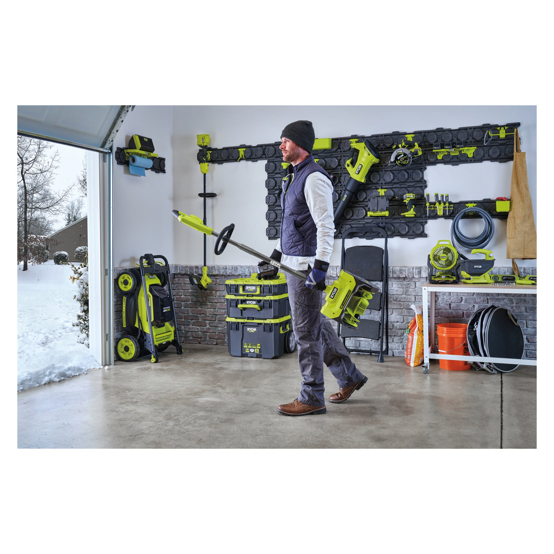 En man i mössa, väst och stövlar går genom ett garage med en Ryobi One+ RY18ST25A-0 trimmer. Gröna och svarta Ryobi-elverktyg och utrustning är prydligt arrangerade på väggar och hyllor. Snö kan ses utomhus genom den öppna garageporten.
