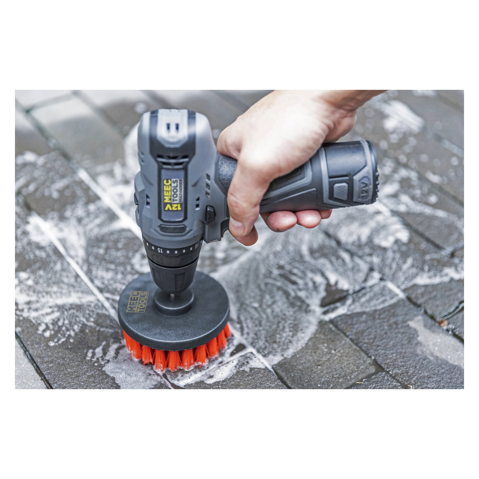 En hand greppar en sladdlös borr utrustad med ett Meec Tools Brush-set, och använder det rödborstade tillbehöret för att skrubba våta, tvåliga utomhusplattor. Skummande tvål täcker en del av ytan.