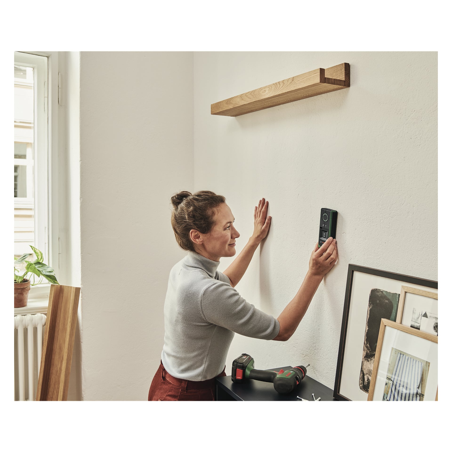 Man använder ett Bosch Truvo digitalt laservattenpass på väggen för att förbereda montering av en trähylla ovanför. I närheten placeras en borr och några inramade fotografier ovanpå ett skåp, med ett fönster och en krukväxt synliga i bakgrunden.