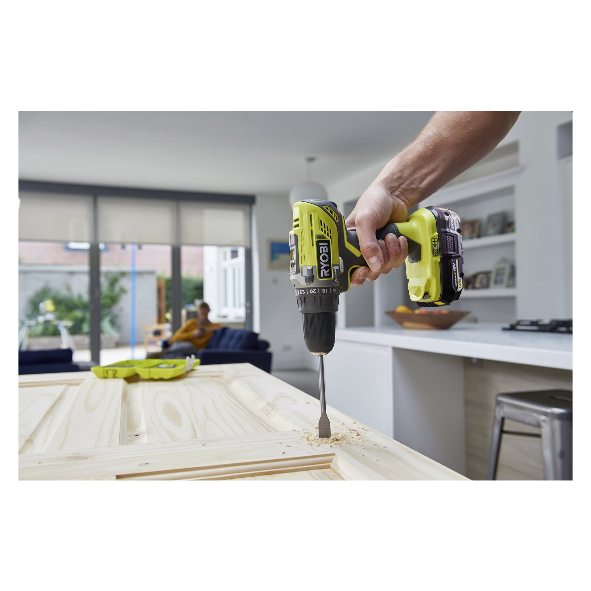 Någon använder en Ryobi One+ skruvmejsel utrustad med ett borrset och batteri R18DD3-1C20GA31J för att borra i en trädörr i ett modernt kök, vilket ger sågspån på ytan. I bakgrunden sitter en annan person i en soffa.