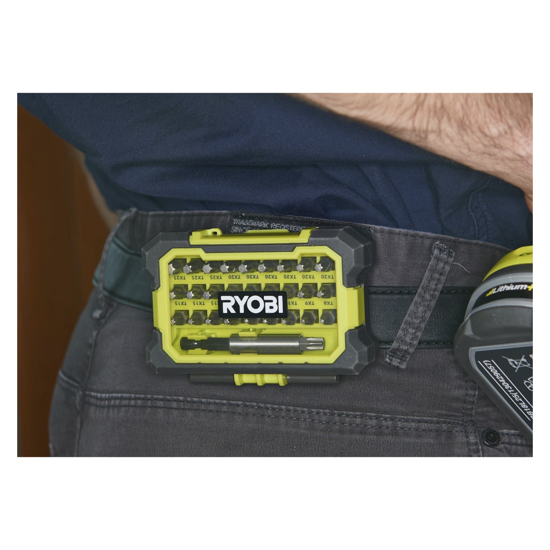 En närbild visar en person med en Ryobi One+ skruvmejsel, biten inställd, och ett R18DD3-1C20GA31J-batteri fäst vid bältet. Det gula och svarta fodralet innehåller flera märkta borr, medan en del av en Ryobi-borr finns vid ramen ytterligare.